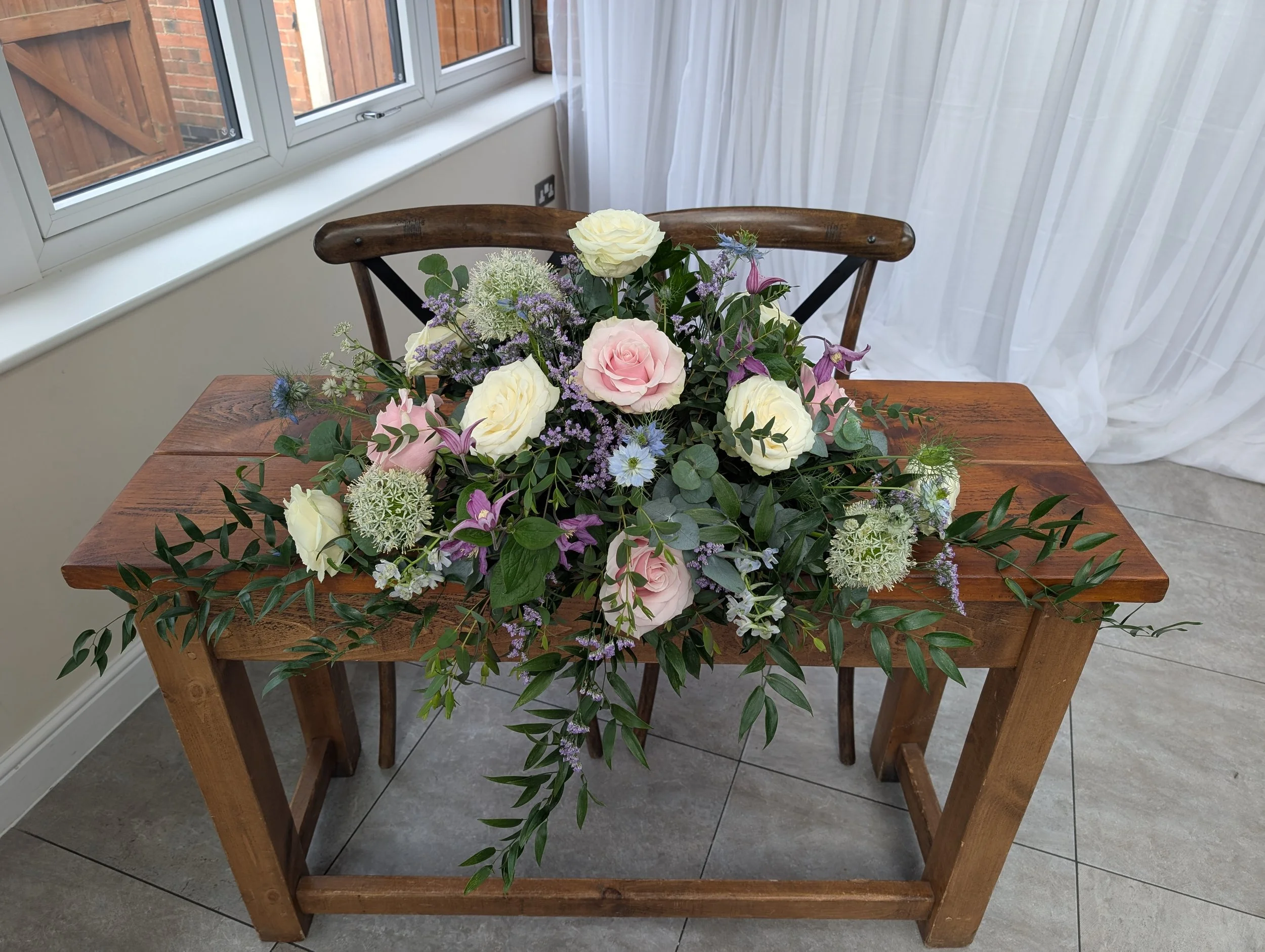 A wooden table with a large floral arrangement of white, pink, and purple flowers with green leaves.