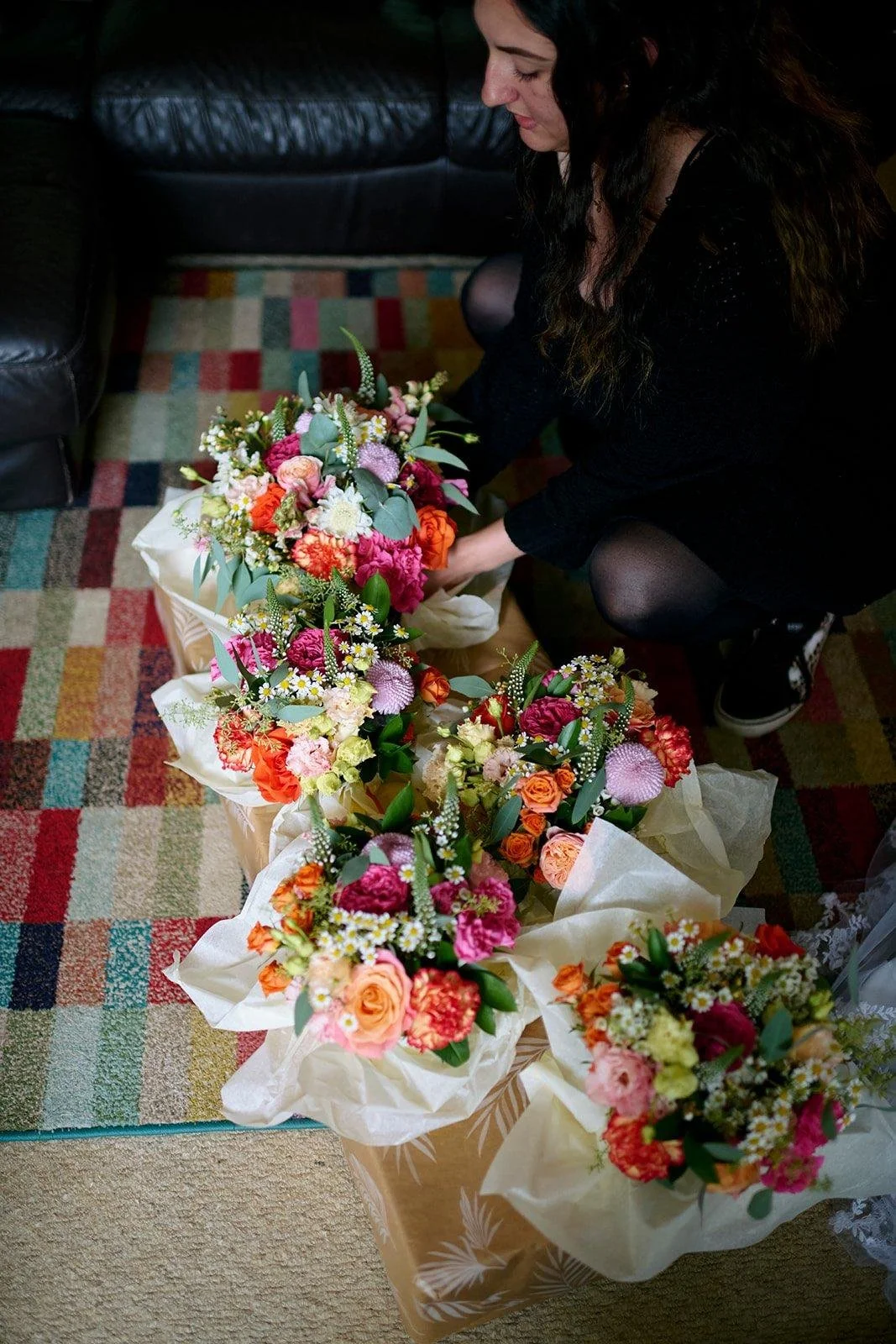 A woman arranging colorful flower bouquets on a table in a living room, with a black couch and a multicolored rug.