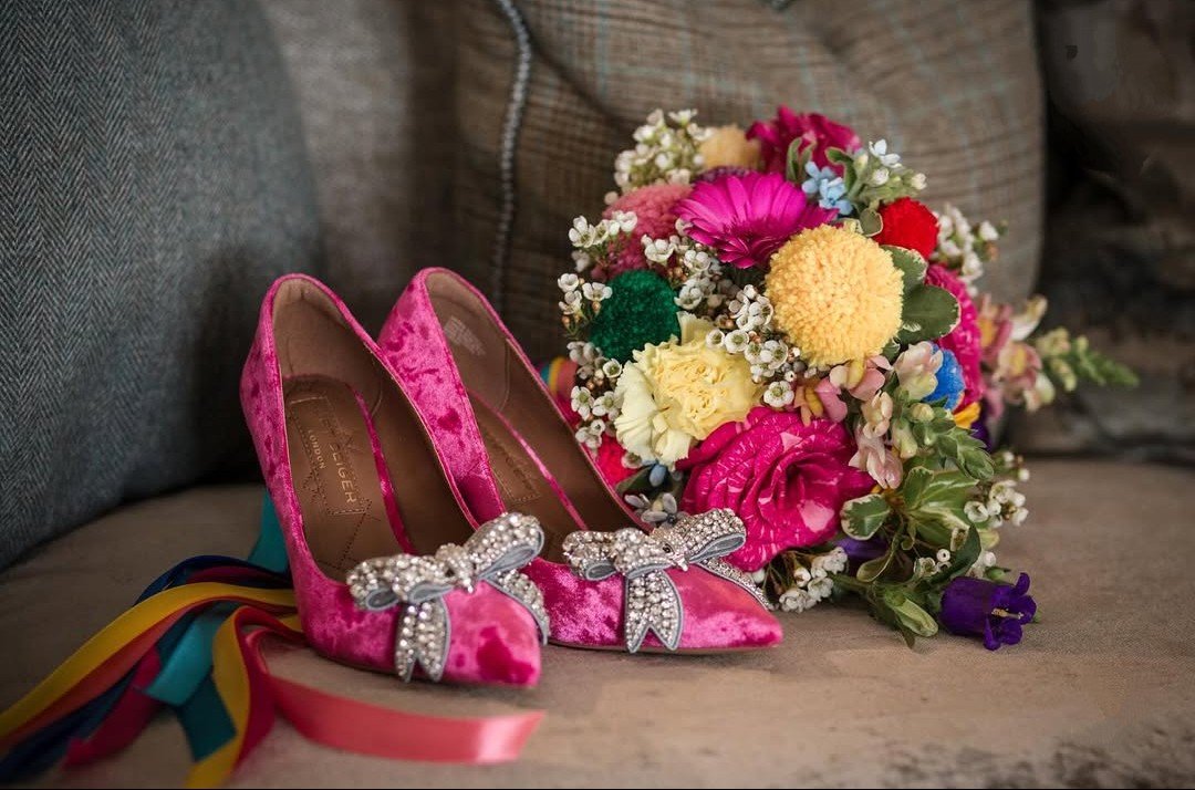 Pink velvet high heels with rhinestone bows next to a colorful flower bouquet on a beige surface.