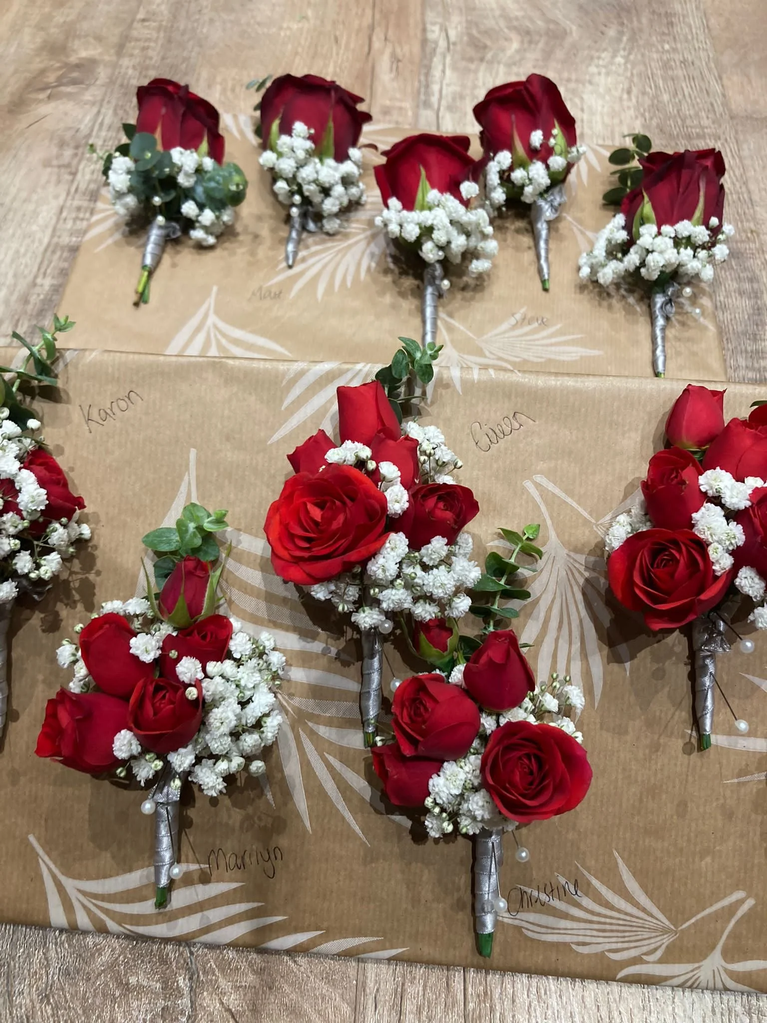 Several corsages made of red roses, baby’s breath, and greenery with wrapped stems decorated with silver and pearl accents, are arranged on a brown paper background with handwritten names.
