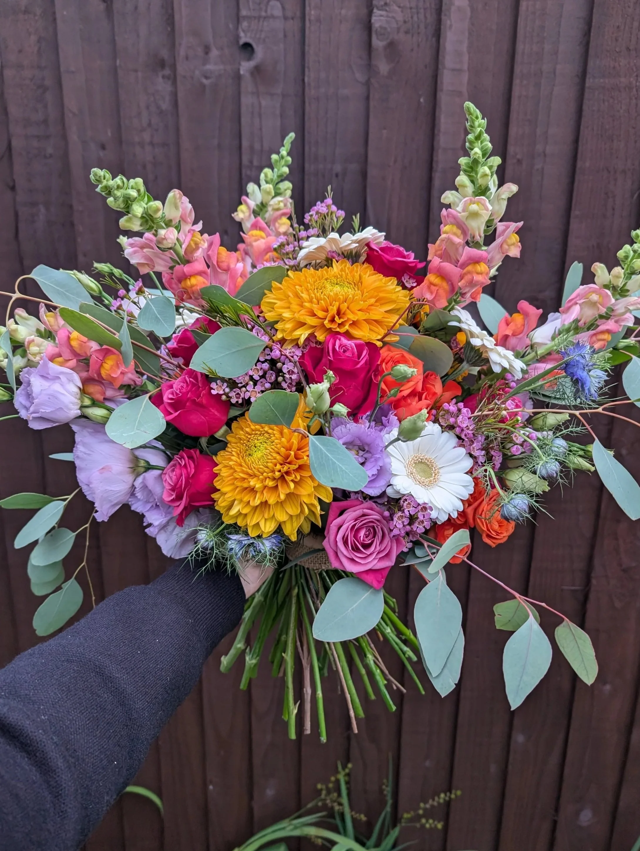 Colorful bouquet of flowers including pink roses, yellow chrysanthemums, white daisies, purple lisianthus, and eucalyptus leaves, held in front of a wooden fence.