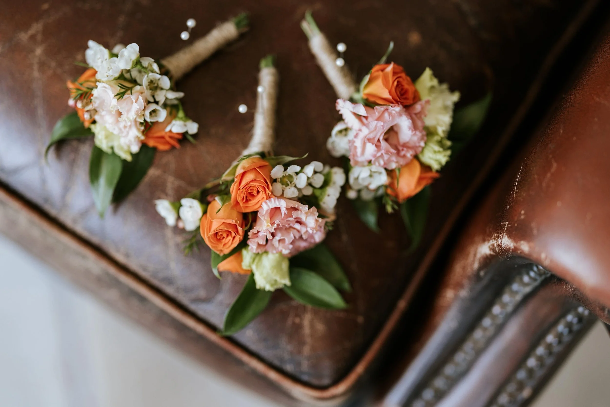 Three boutonniere floral arrangements with orange roses, pink carnations, white lilies of the valley, and greenery, resting on a wooden surface.
