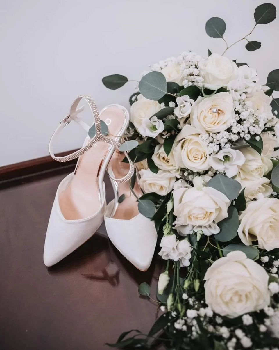 A pair of white high-heeled shoes with ankle straps adorned with rhinestones, placed next to a bouquet of white roses, baby's breath, and eucalyptus leaves on a dark wood surface.