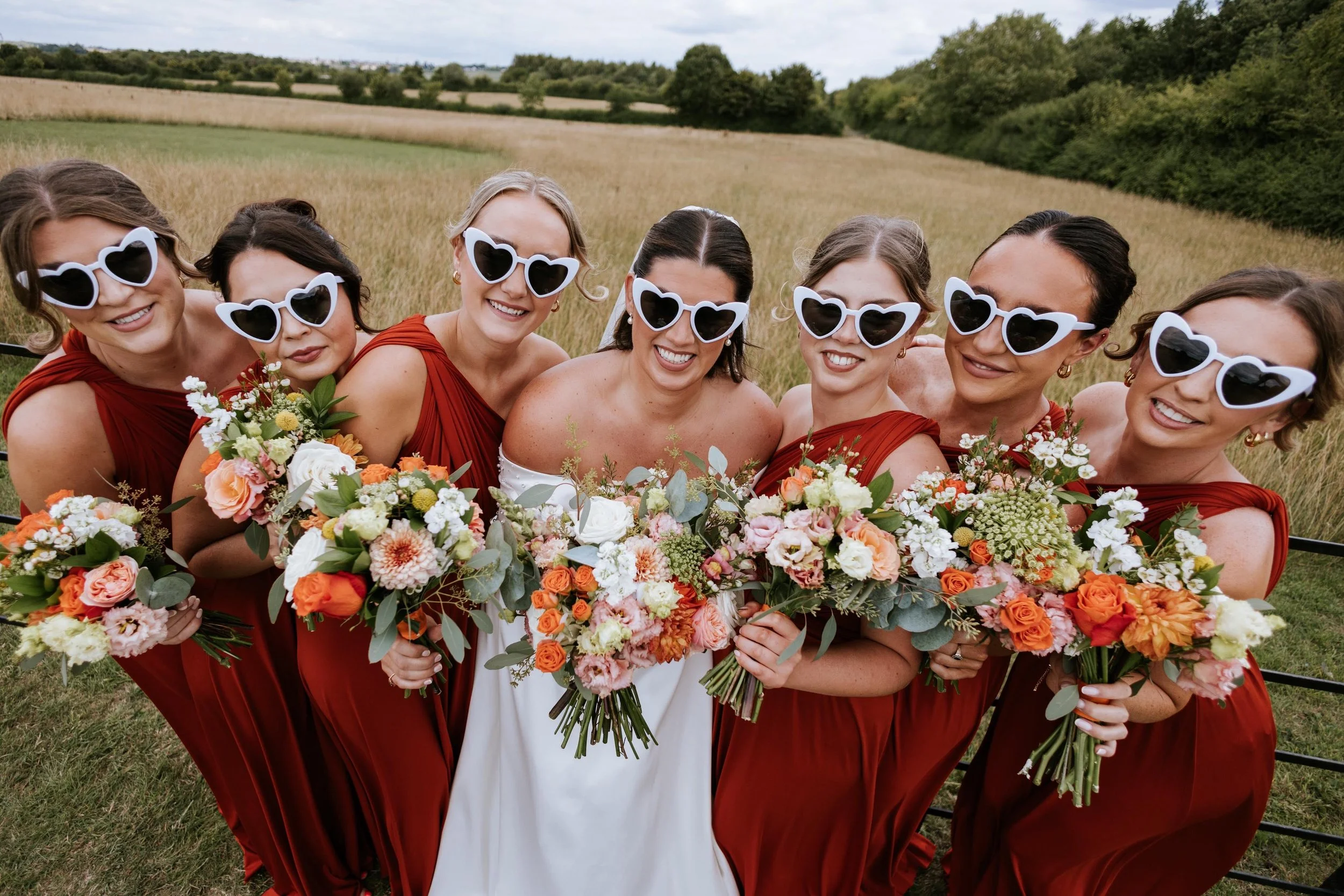 Group of women at a wedding standing in a field, wearing matching red dresses and white heart-shaped sunglasses, holding bouquets of colorful flowers.