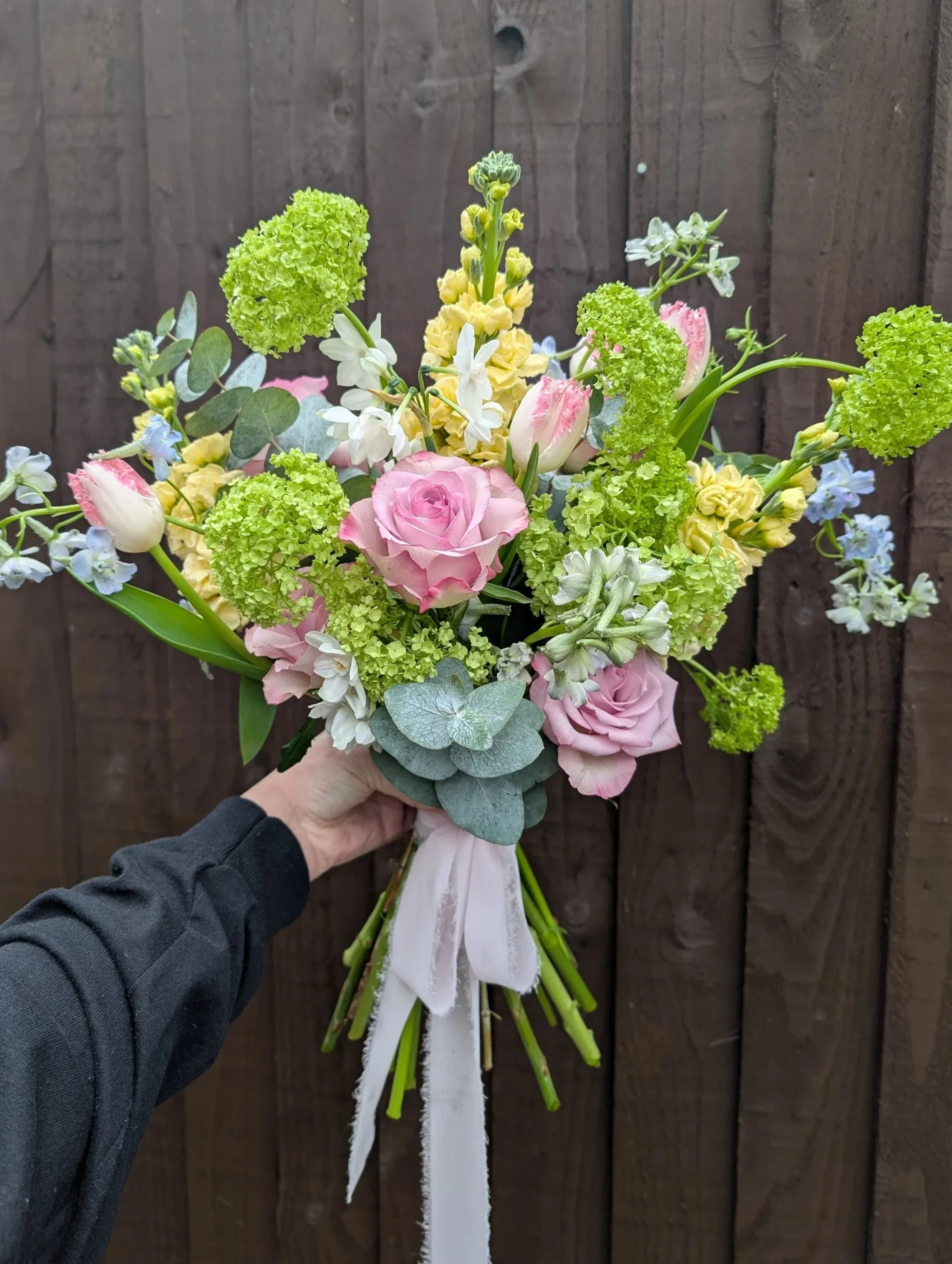A colorful bouquet of pink roses, green hydrangeas, white and pink tulips, and other flowers held by a person in a black long-sleeve shirt against a wooden fence background.