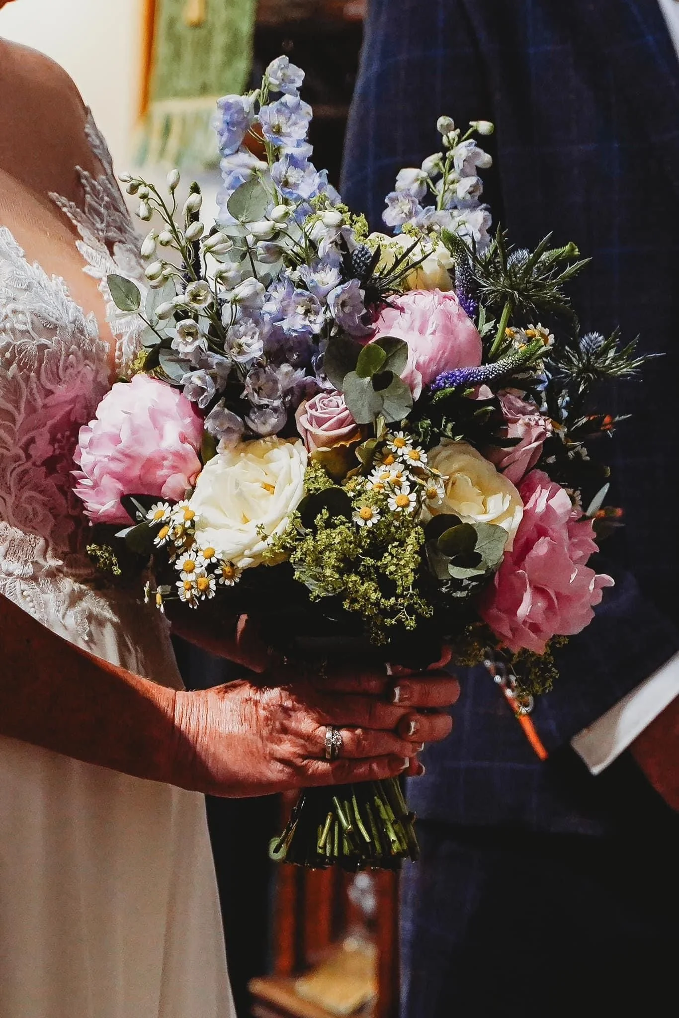 Close-up of a woman holding a bouquet of pink roses, white peonies, blue delphiniums, chamomile, and other mixed flowers.