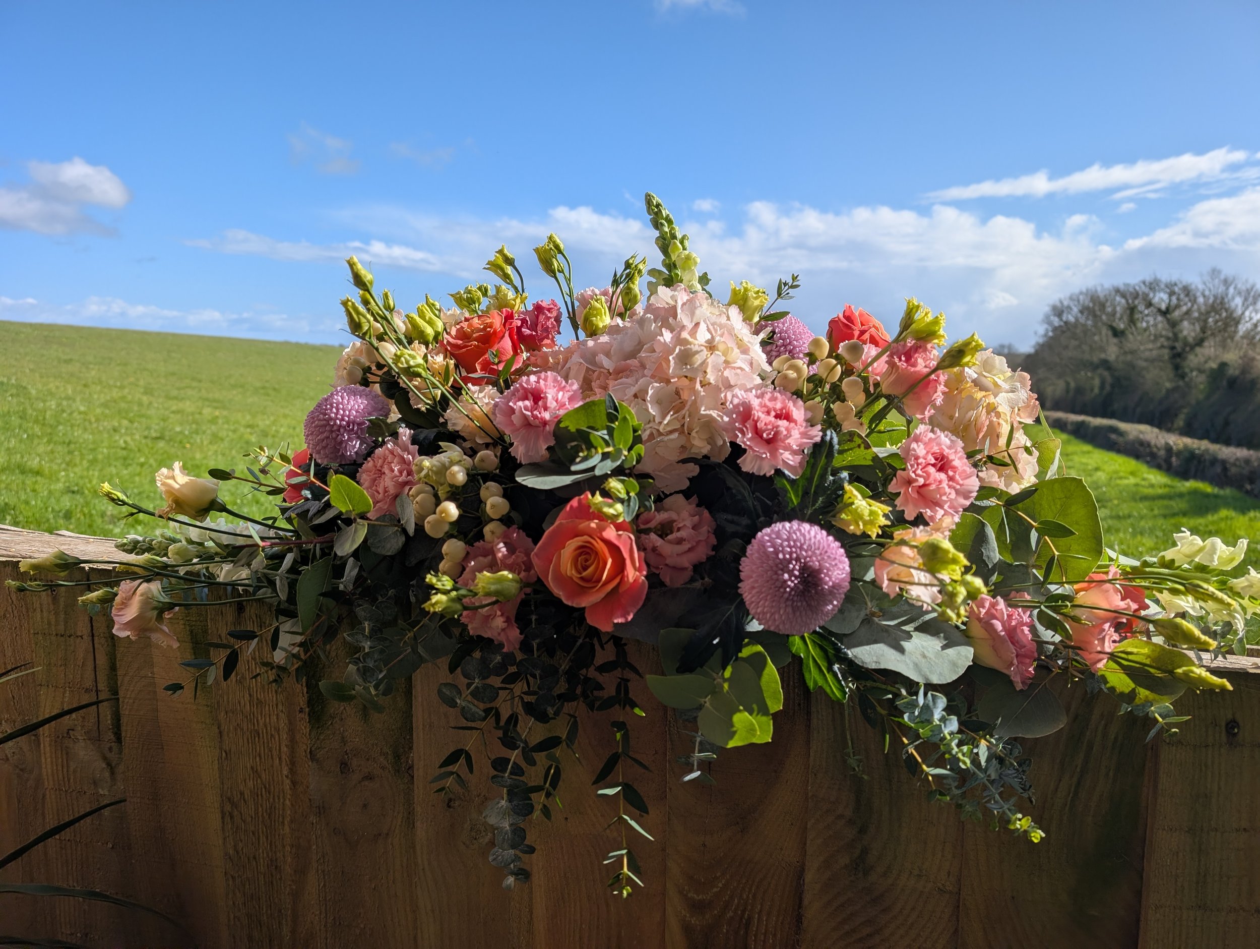 A colorful flower arrangement with roses, carnations, and other flowers resting on a wooden fence outdoors, set against a grassy field and a blue sky with scattered clouds.