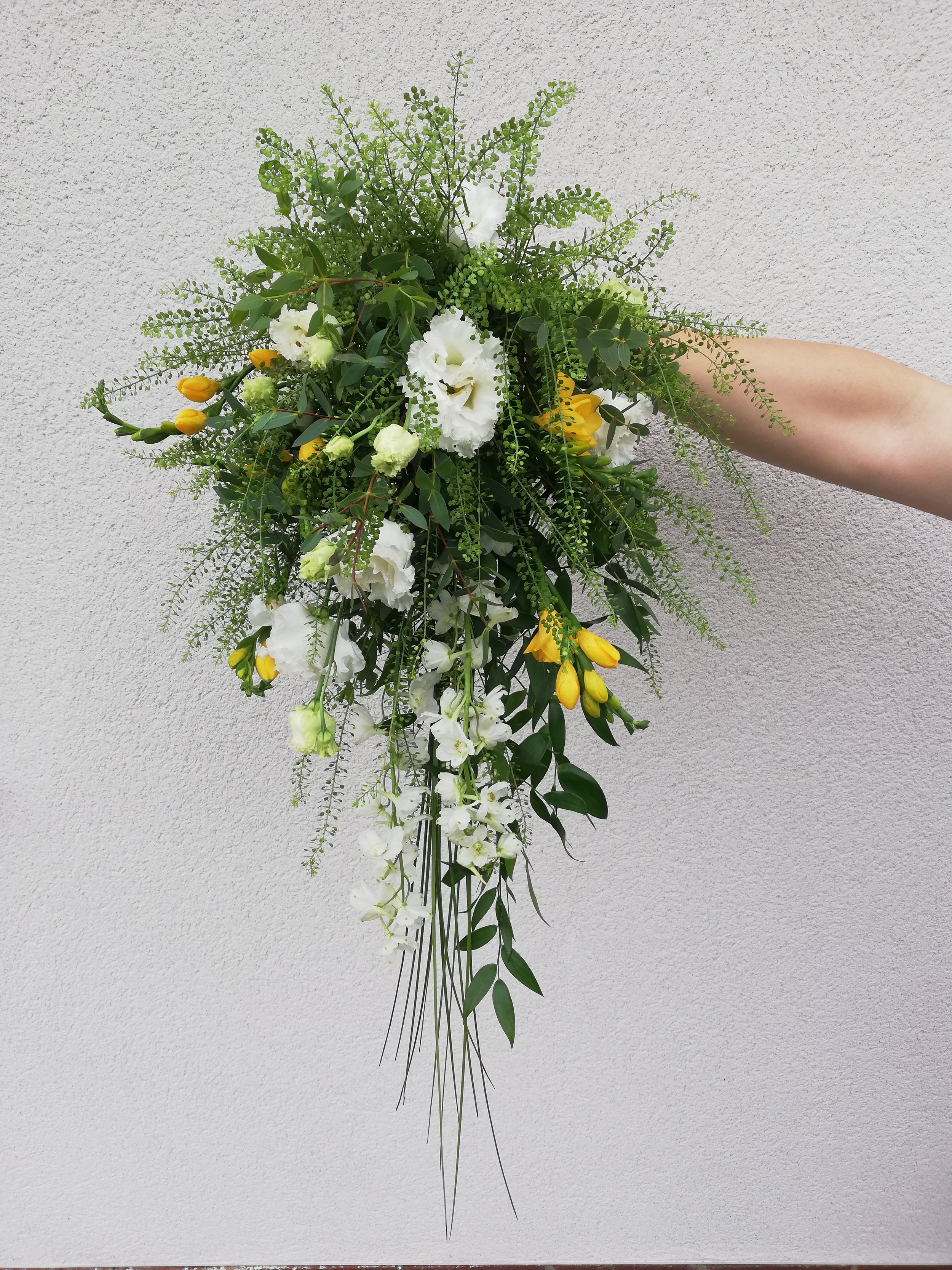 A cascading bridal bouquet featuring white and yellow flowers with green foliage, held by a person's hand against a plain textured background.