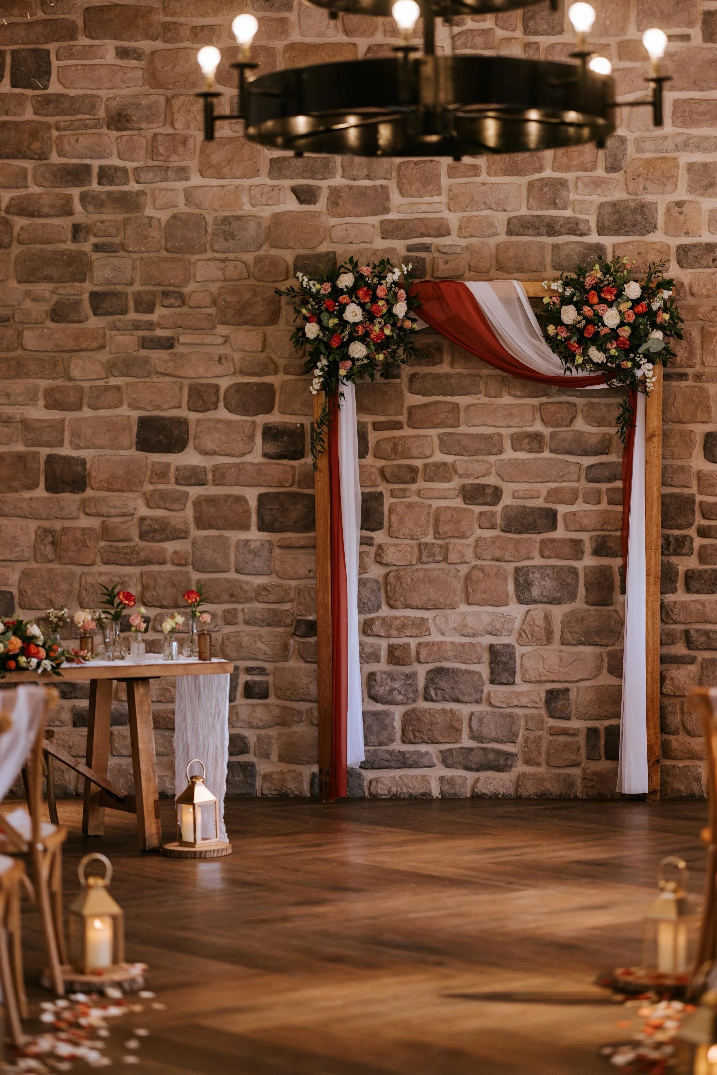 A decorated wedding ceremony space with a rustic brick wall, a floral arch with white, pink, and red flowers, draped with white and red fabric, a wooden table adorned with floral arrangements, candles, and a lantern on the floor.