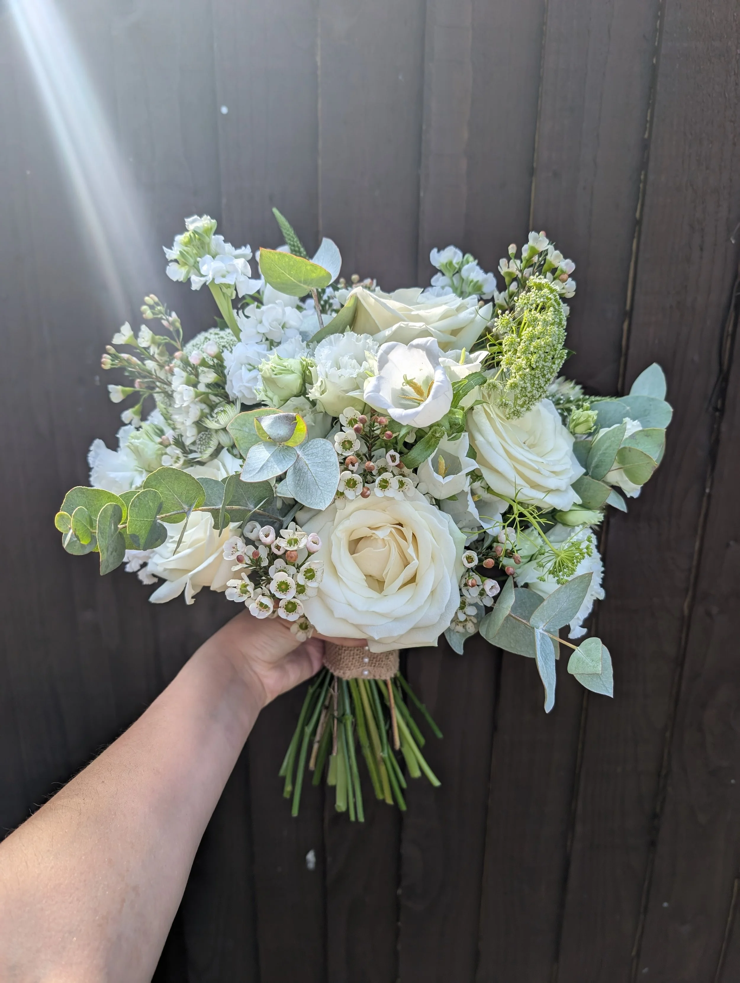 A hand holding a bouquet of white and cream flowers with green leaves and a dark wooden background.