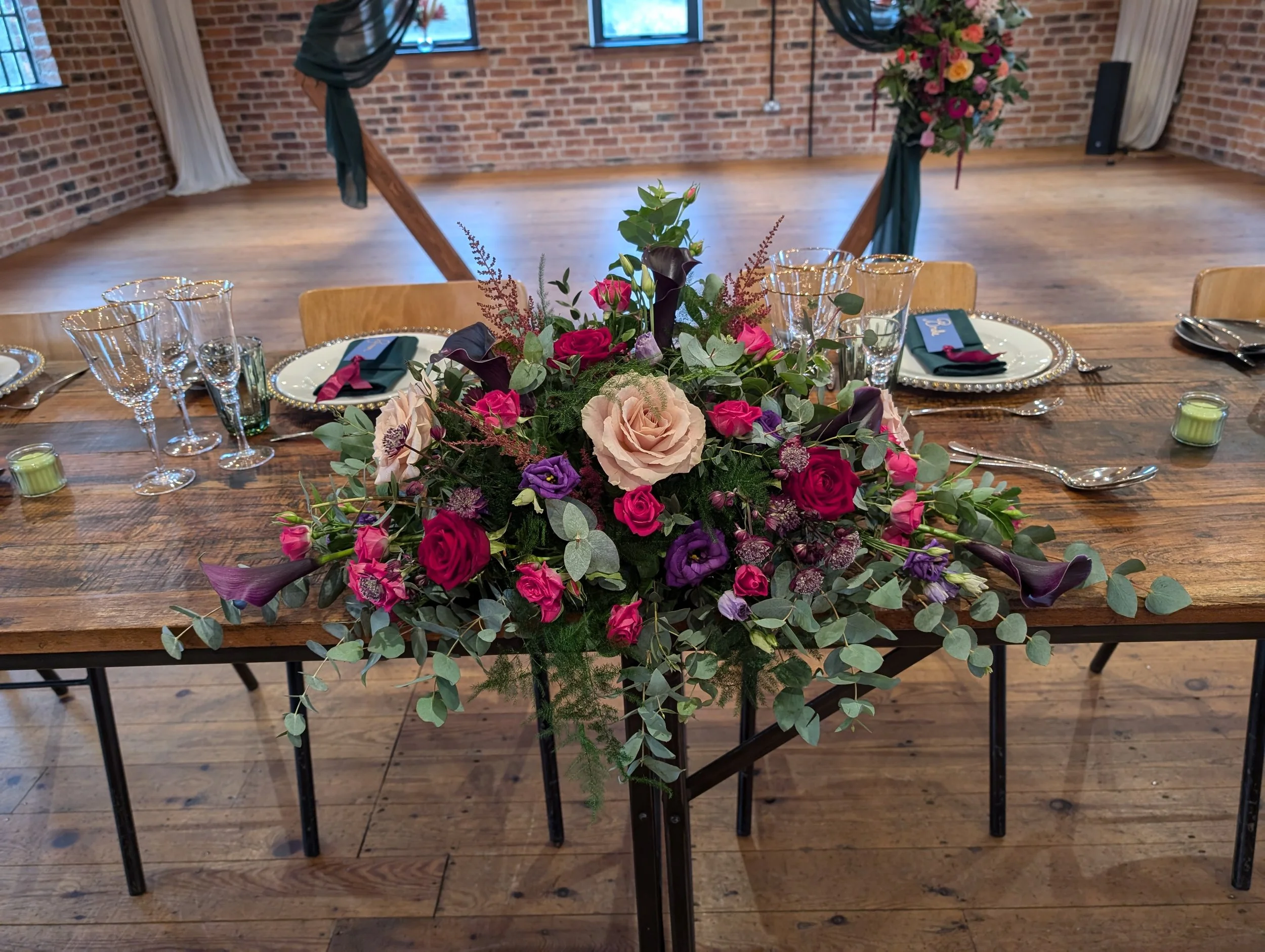 A banquet table decorated with a large colorful floral centerpiece featuring pink, purple, and beige roses, with elegant place settings including plates, glasses, and napkins, in a rustic brick-walled room with wooden floors and a wooden backdrop decorated with green fabric and pink flowers.