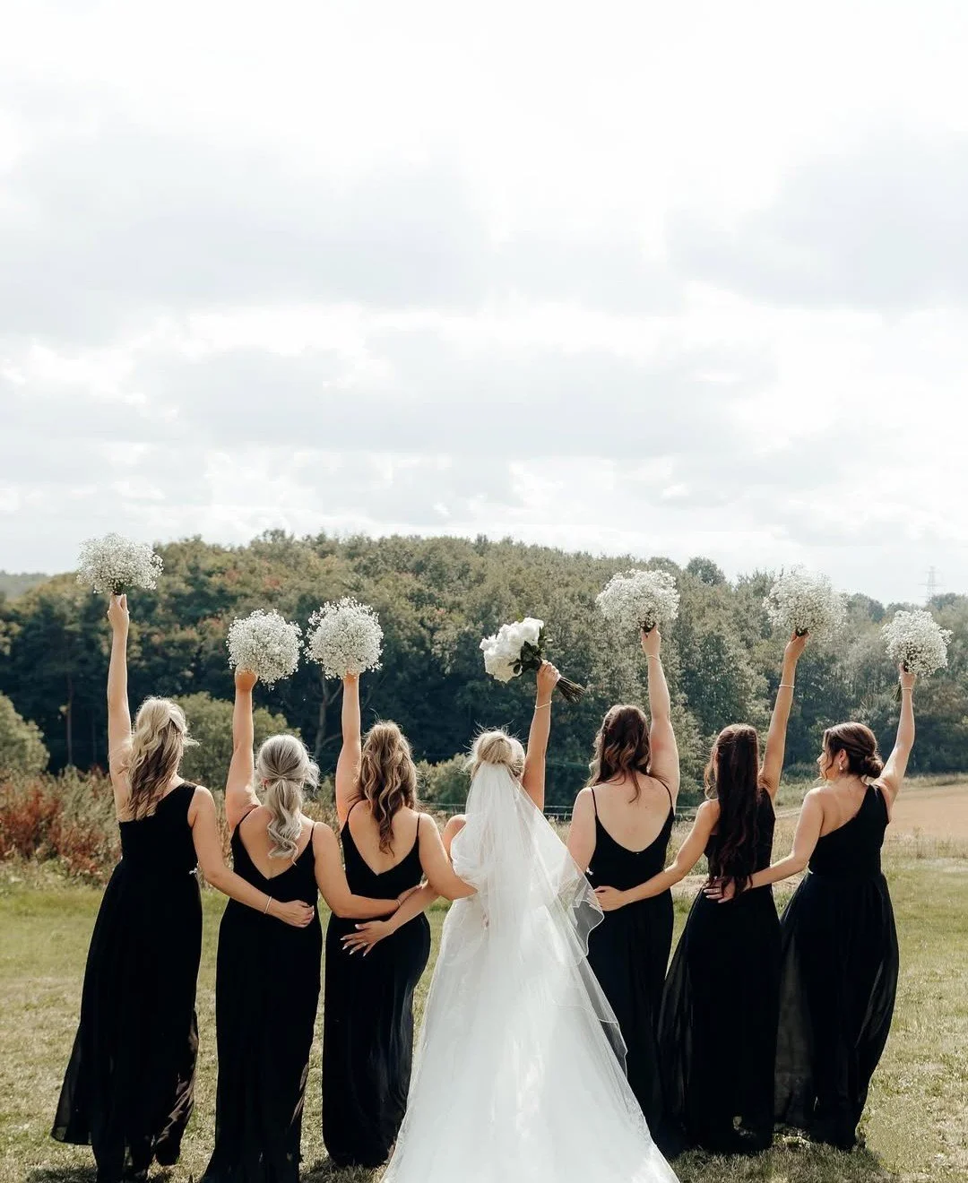Bride in a white wedding dress walking with six bridesmaids in black dresses holding bouquets of white flowers in an open field.