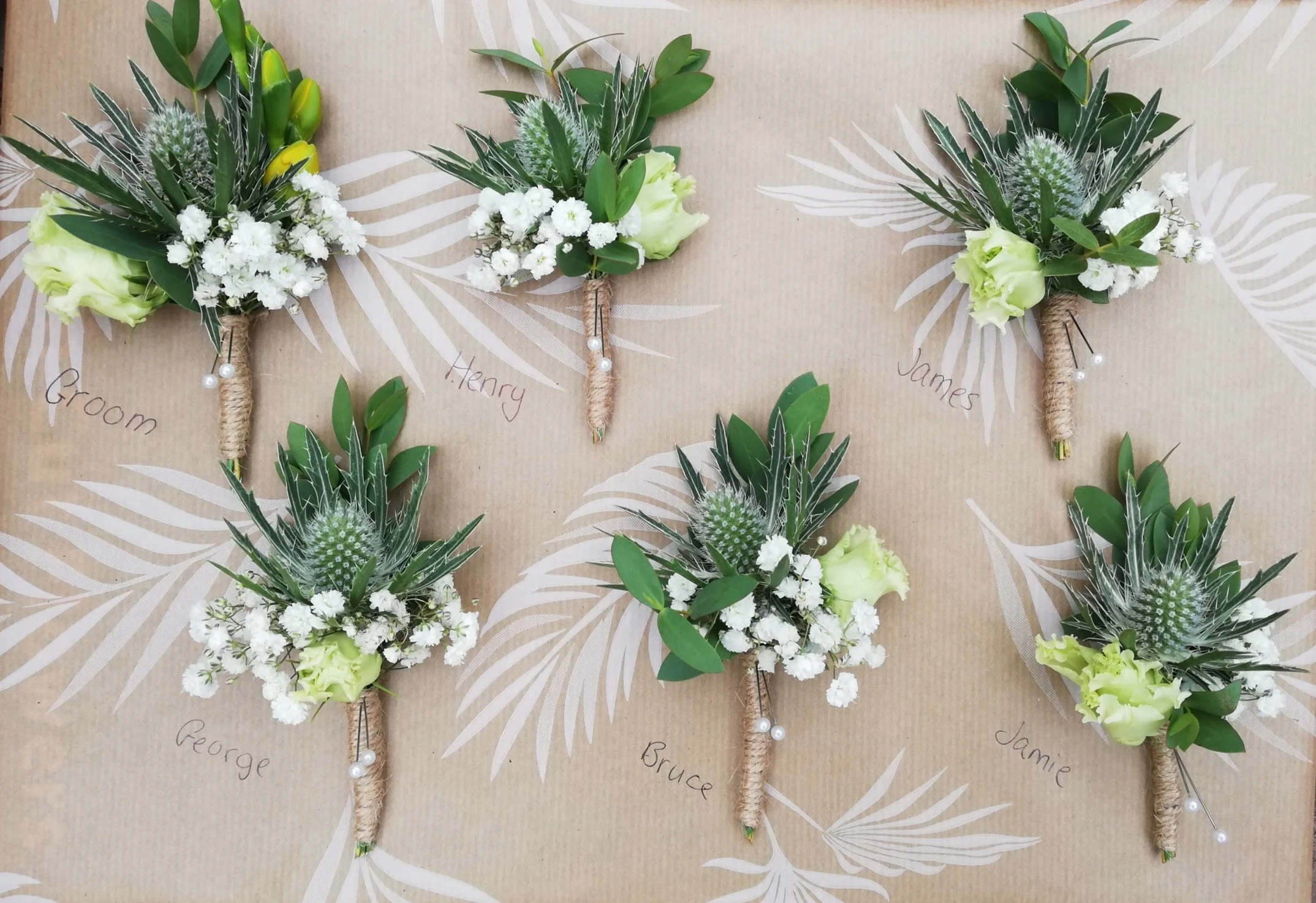 Six boutonniere corsages adorned with white flowers, greenery, and pearl accents, each labeled with names such as Groom, Henry, James, George, Bruce, and Jamie, laid out on a beige background with white leaf patterns.
