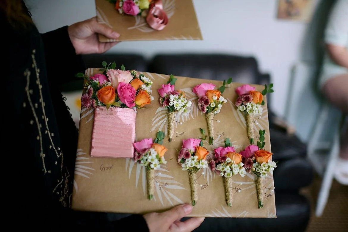 Person holding a wrapped gift box with pink, orange, and white floral corsages and boutonnieres for a wedding, labeled with roles such as groom, bride, and others, in a room with a blurred background.