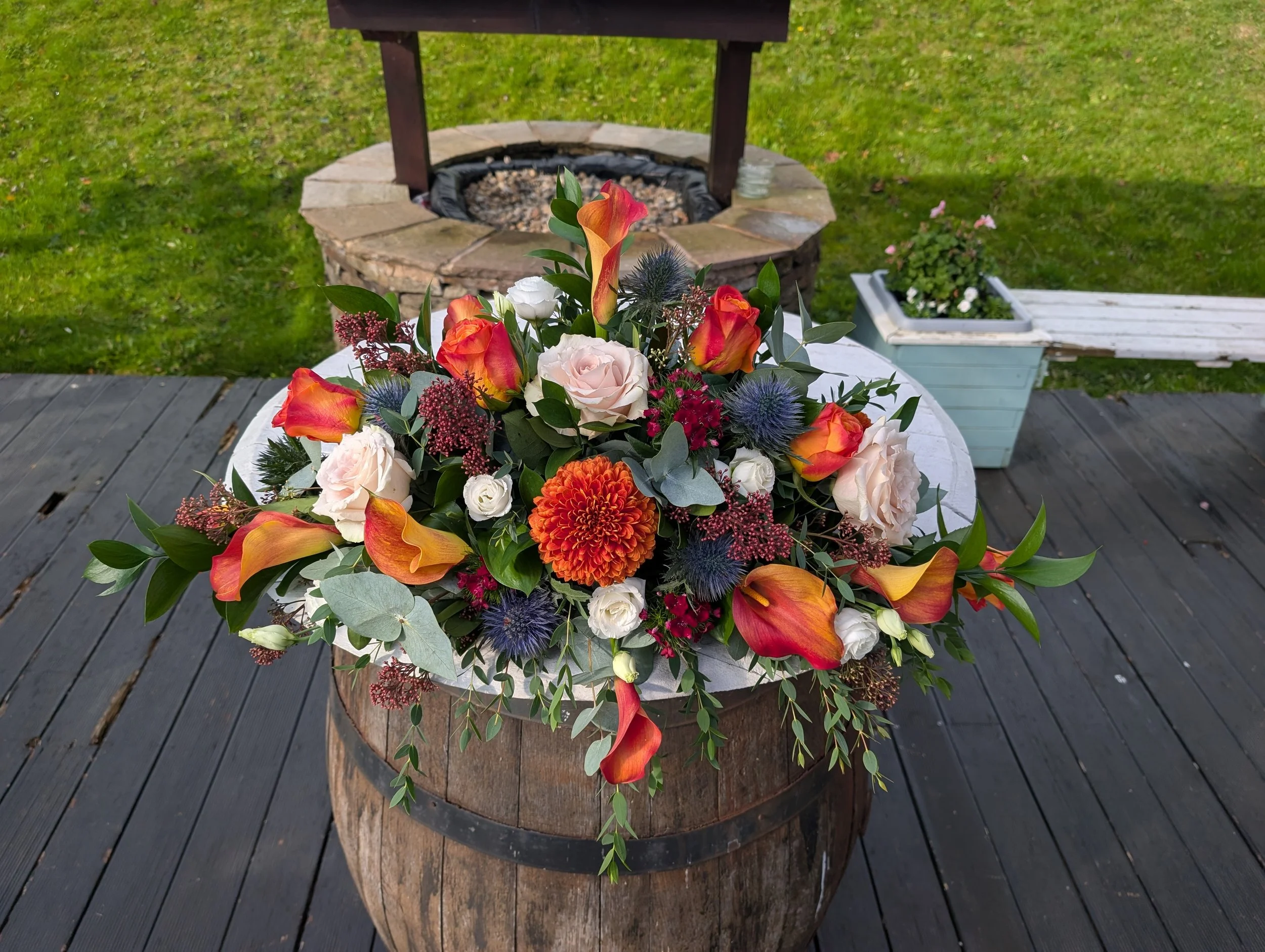Colorful flower arrangement with roses, calla lilies, dahlias, thistles, and greenery in a wooden planter on a wooden deck.