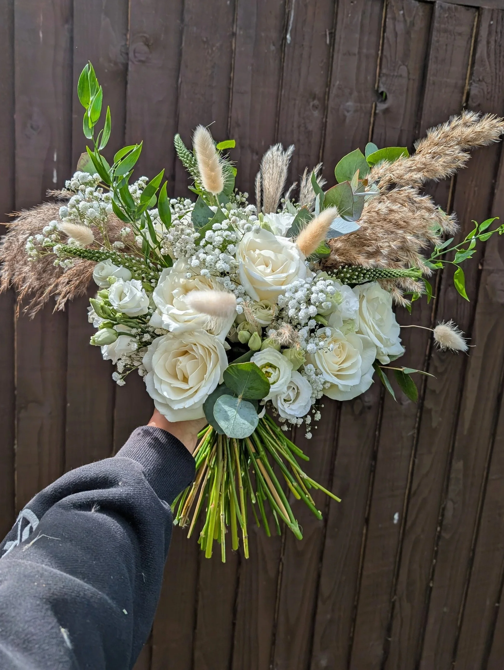 Hand holding a bouquet of white roses, baby's breath, and green foliage against a wooden fence.