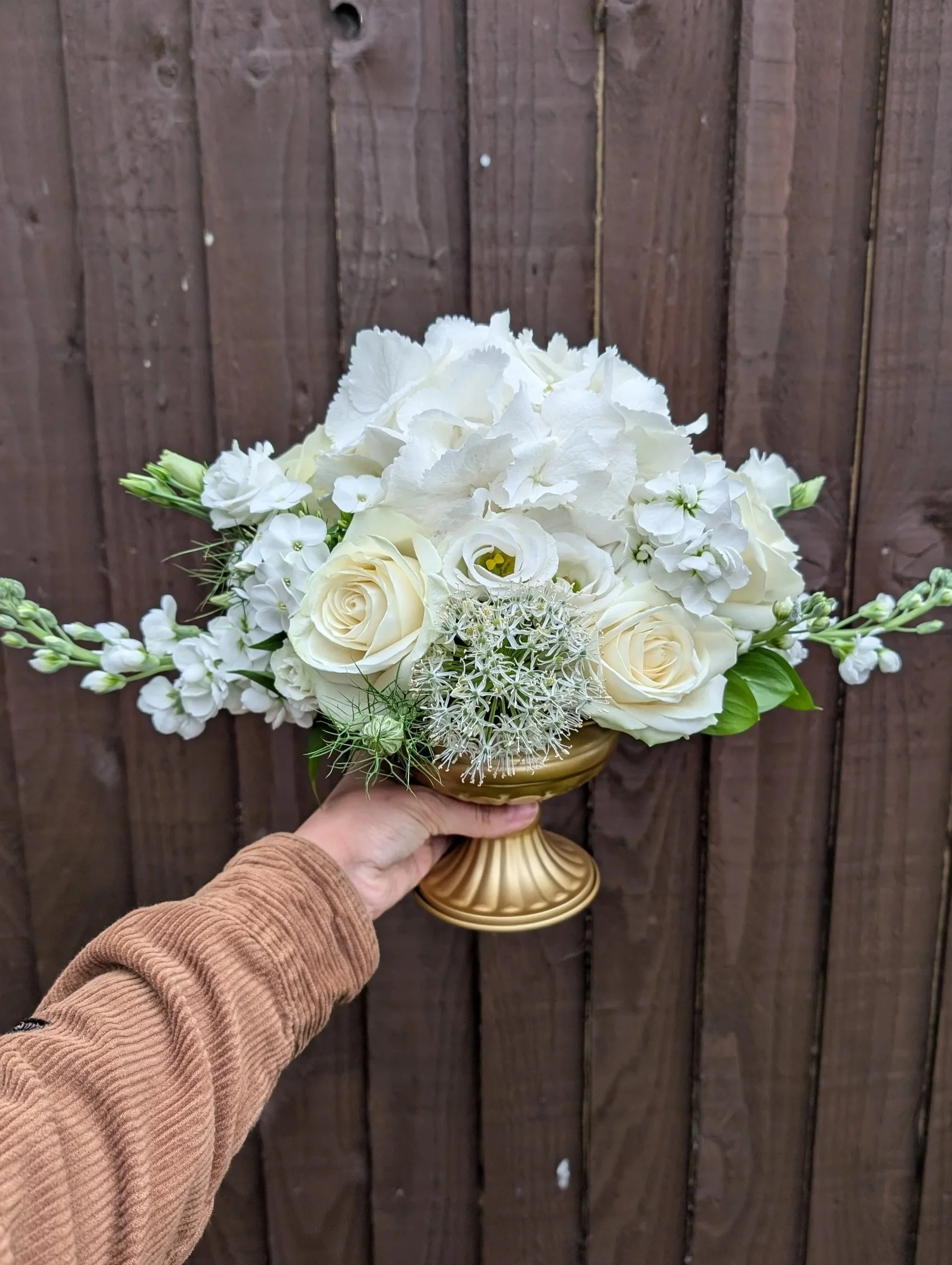 Hand holding a gold-colored vase filled with white flowers, including roses and hydrangeas, against a wooden fence background.