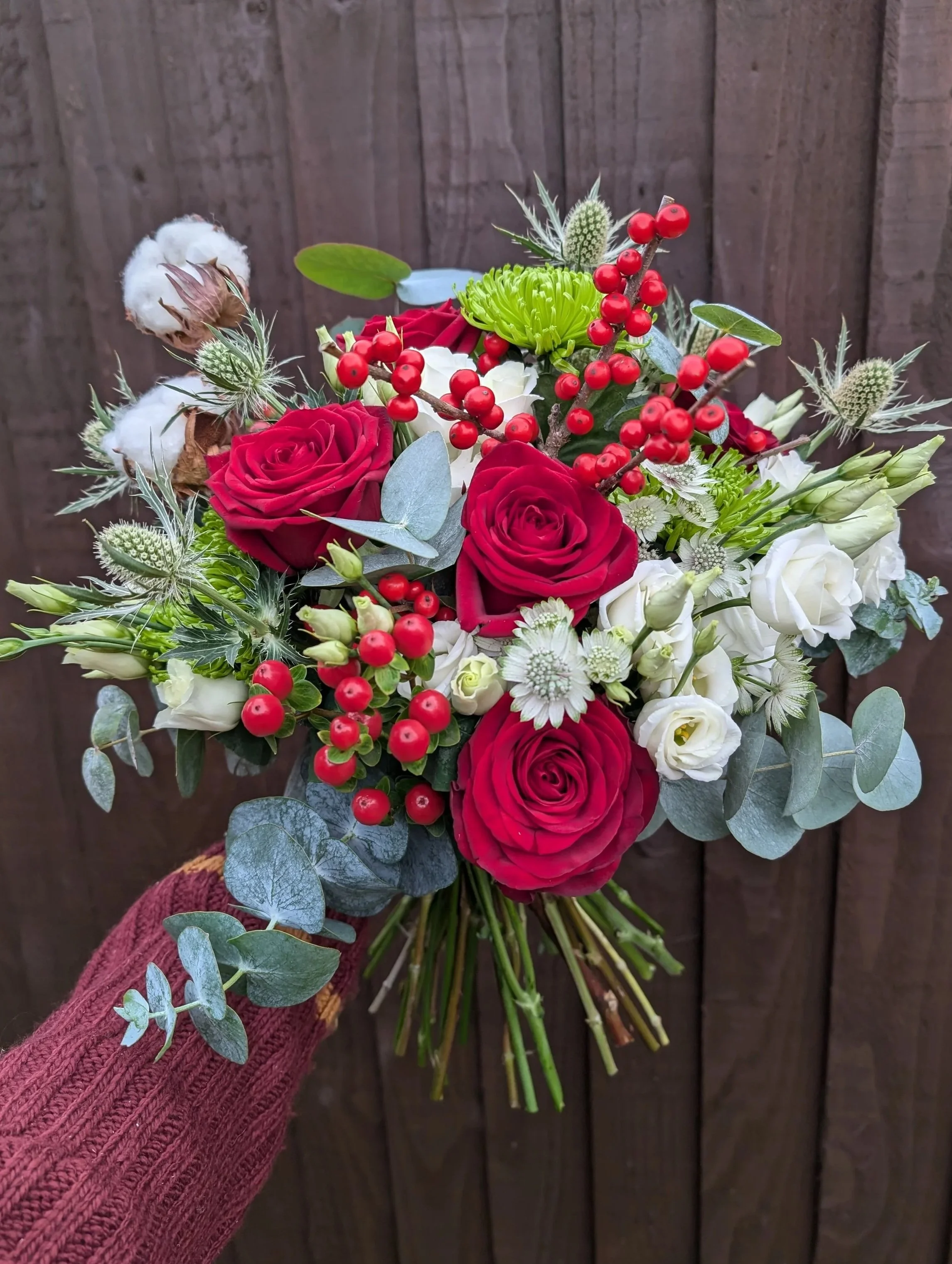 A hand holding a bouquet with red roses, white flowers, red berries, green chrysanthemums, and eucalyptus against a wooden background.