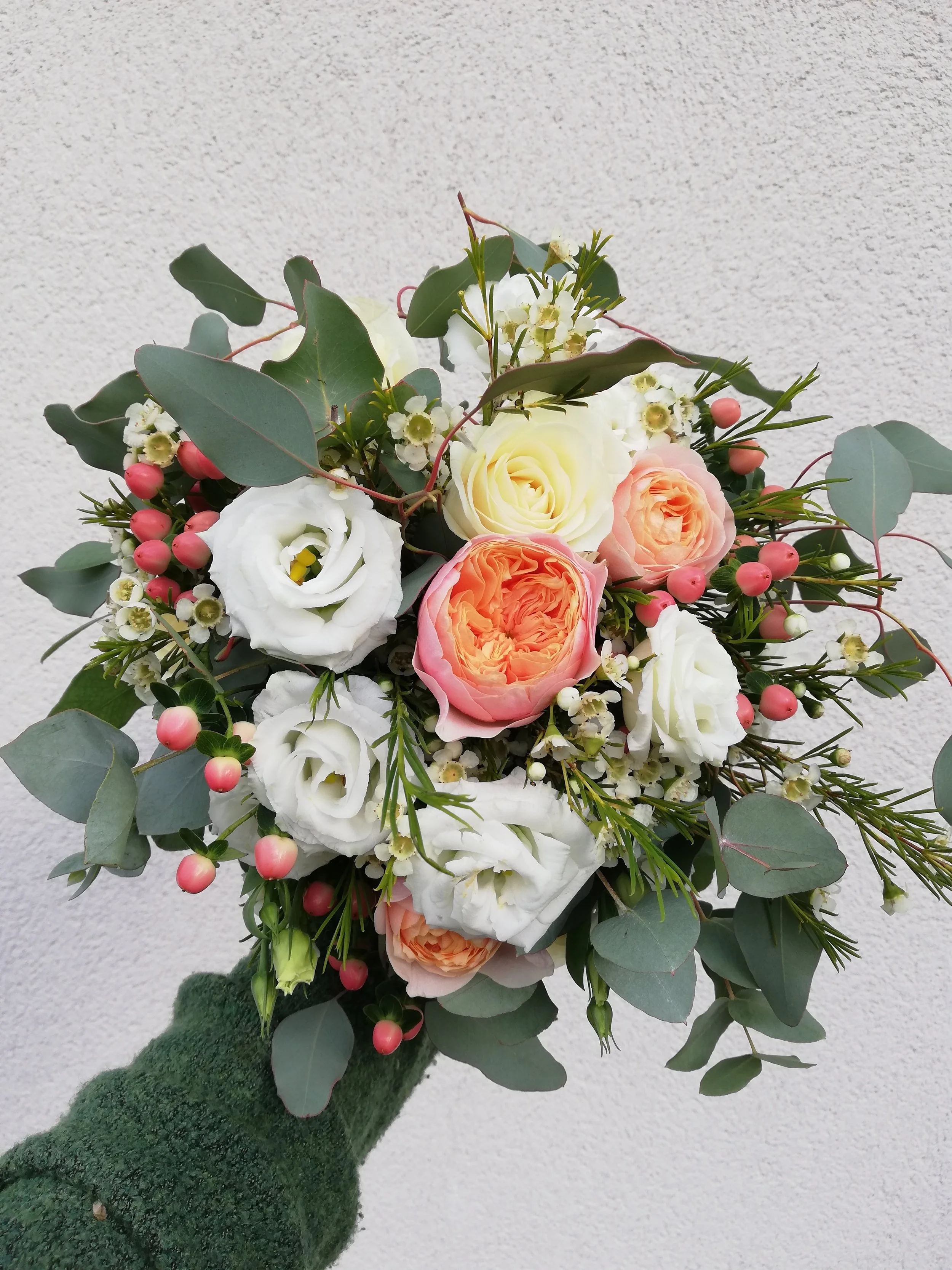 A hand holding a bouquet of mixed flowers including white roses, pink roses, small white flowers, and eucalyptus leaves against a white textured wall.
