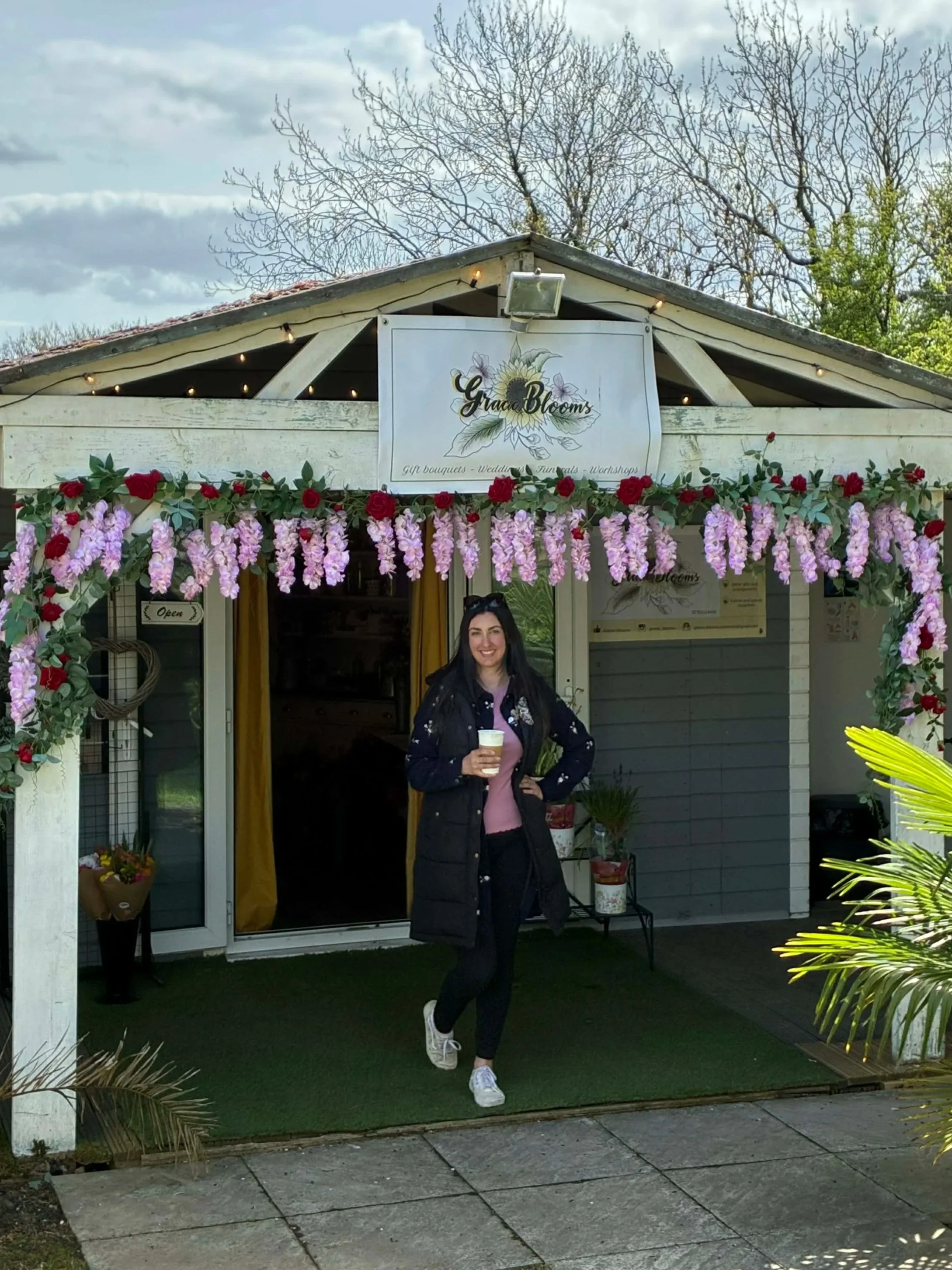 A proud florist business-owner stood in front of her shop in Clay Cross. It is decorated with pink flowers and green foliage, and the sign shows the shop's logo and name, "Gracie Blooms".