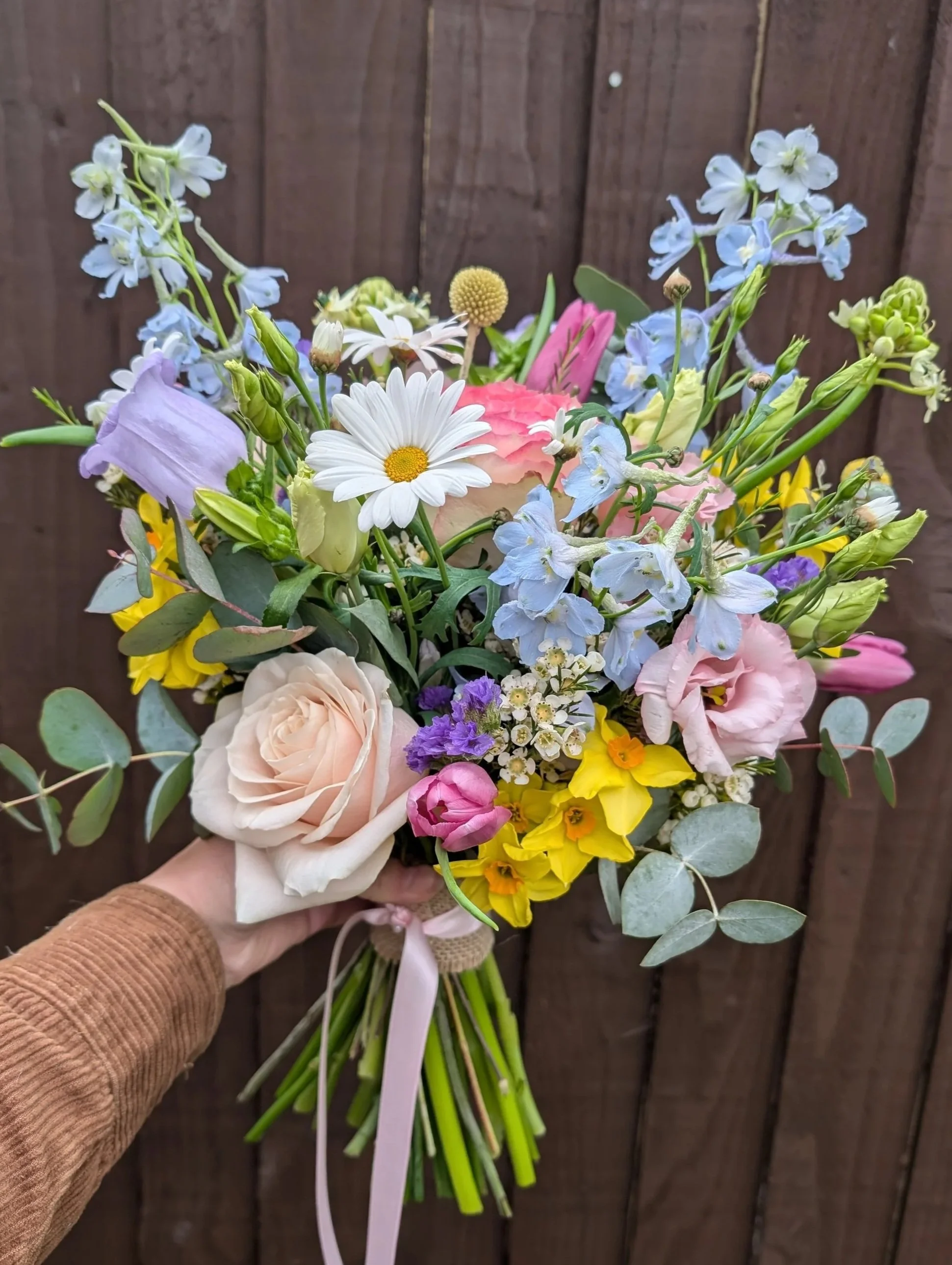 A hand holding a colorful bouquet of mixed flowers including roses, daisies, delphiniums, and other blooms against a wooden background.