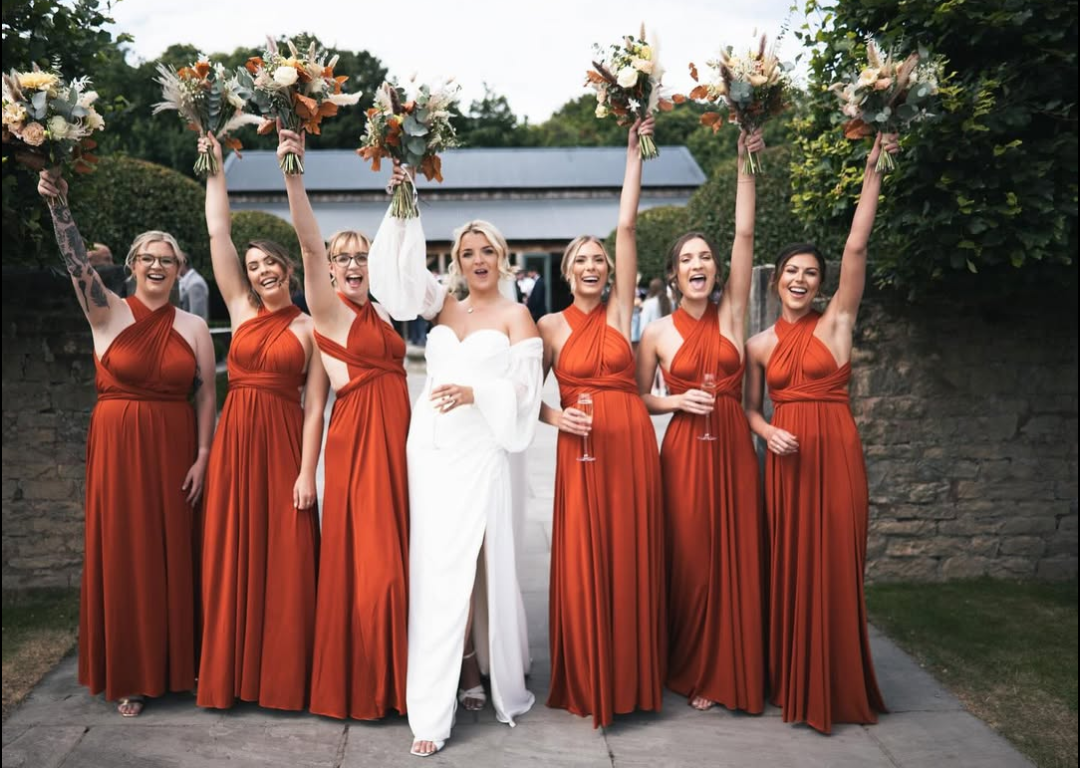Group of bridesmaids in burnt orange dresses celebrating with a bride in white at an outdoor wedding reception.