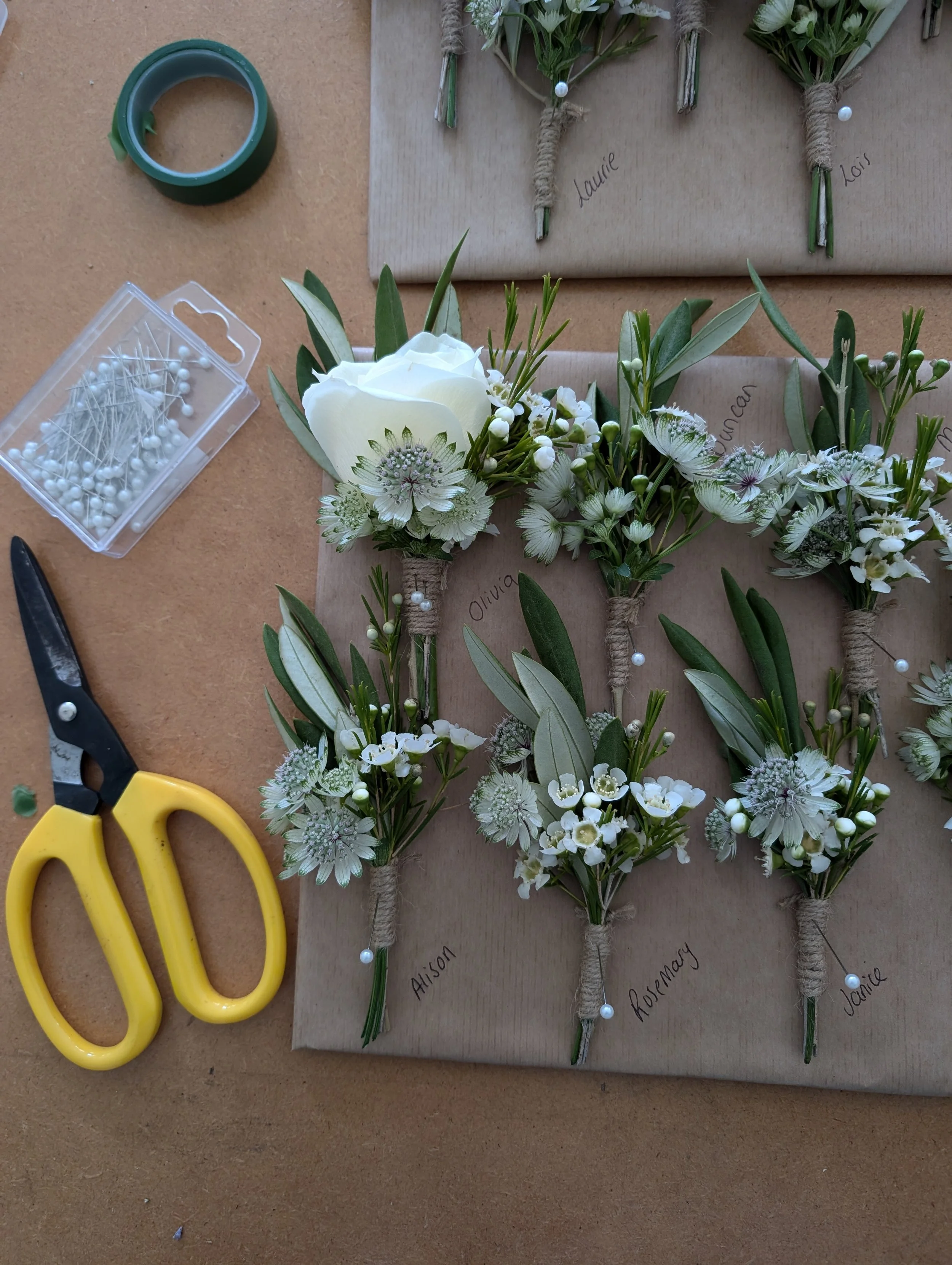 Hands-on image of boutonnières being assembled with white flowers, green leaves, and labeled with names, on brown paper with scissors, pins, and floral tape nearby.