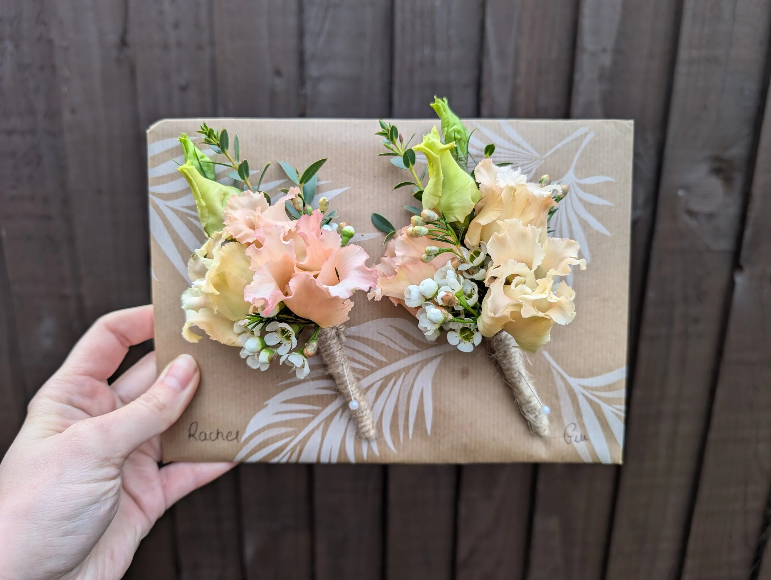 Hand holding a wrapped gift with floral boutonnières attached, on brown paper with white leaf print, background of wooden fence.