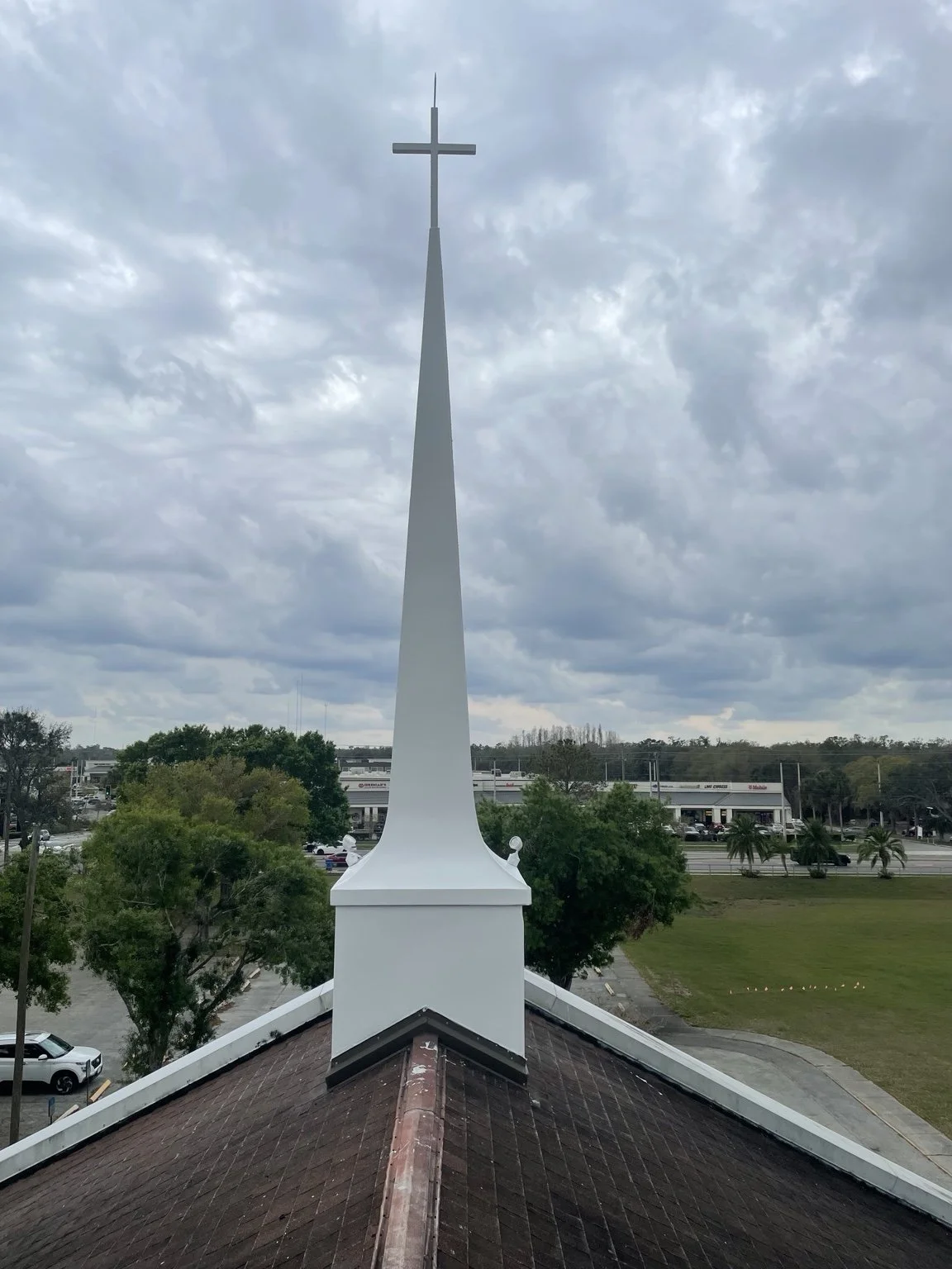 White church steeple with a cross, viewed from the roof with trees and a parking lot below.