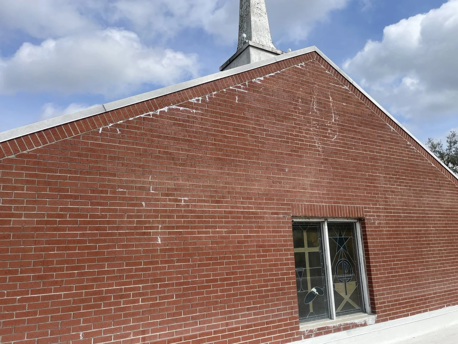 Red brick church exterior with a stained-glass window, white steeple, and blue sky.