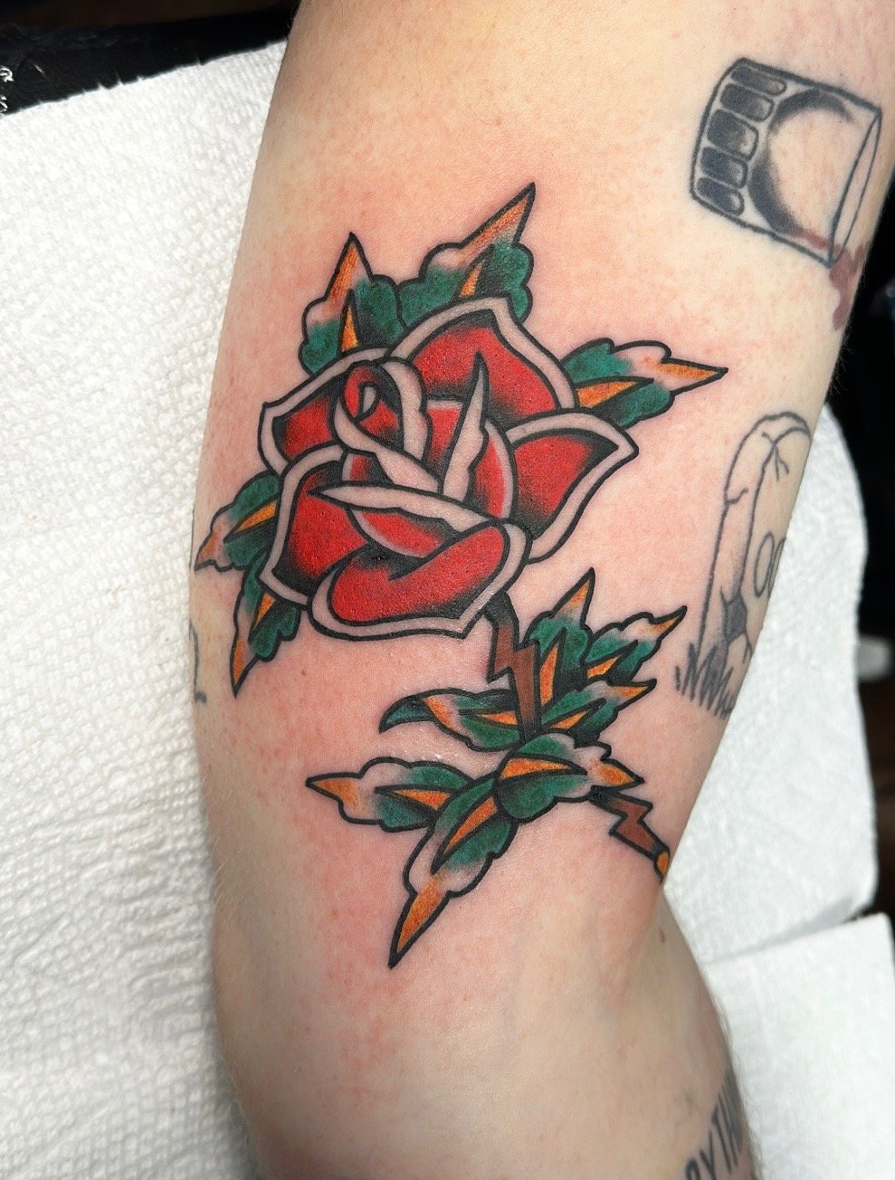 Tattoo featuring a traditional style red rose with green leaves and orange accents on an arm.