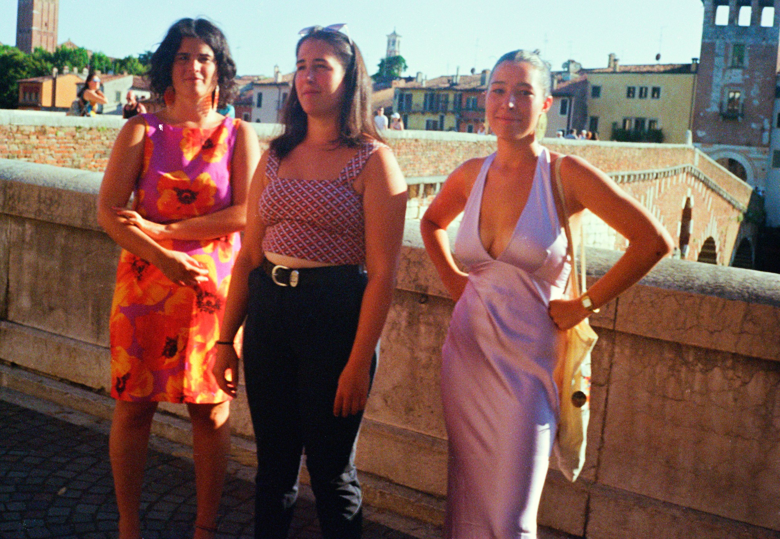 Three women standing outdoors in a sunny, urban setting with historical buildings and a bridge in the background.
