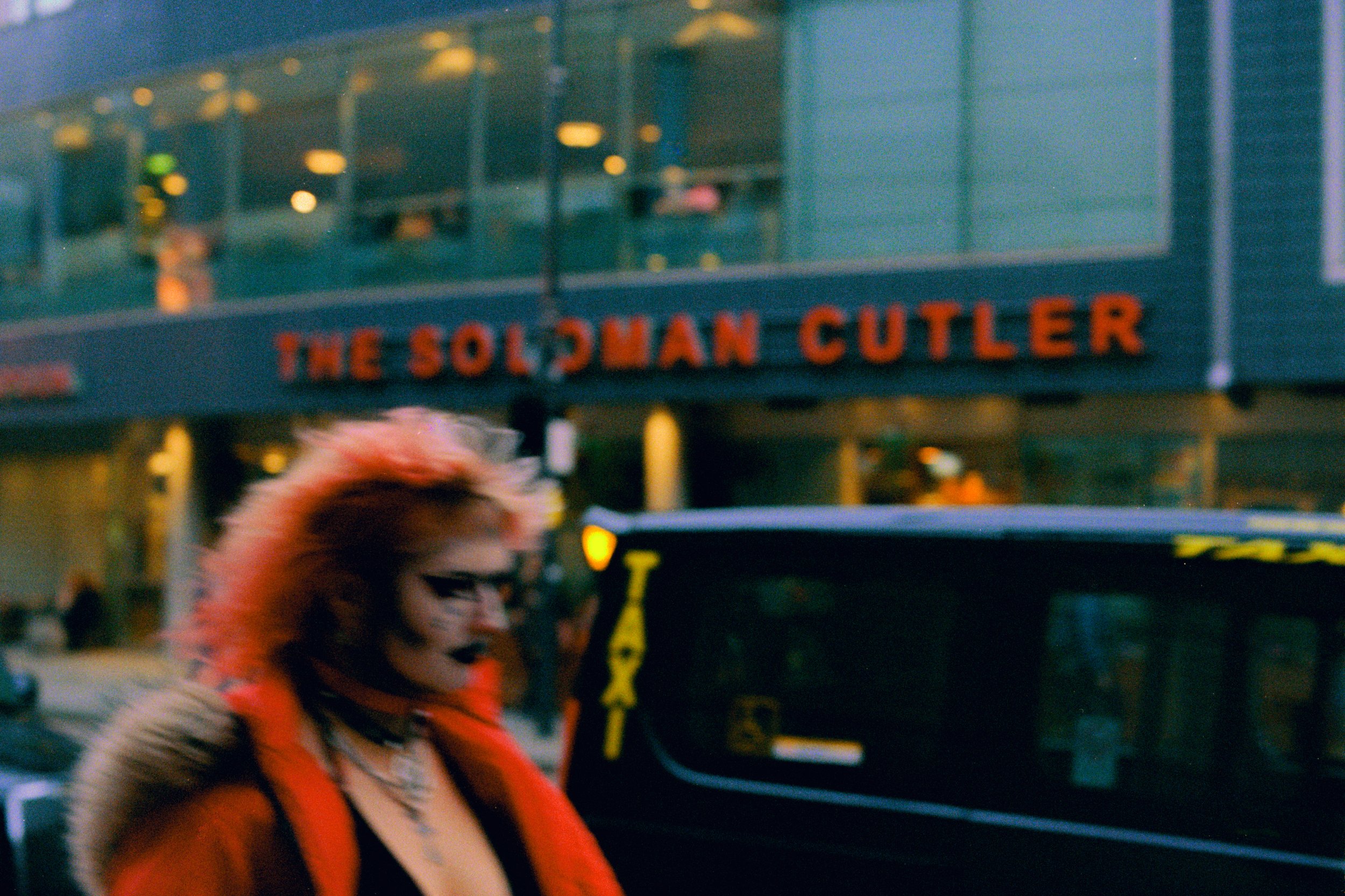 A person with vibrant, pink and orange hair styled in an edgy, messy manner, wearing dark makeup with bold eyeliner, black lipstick, and a black choker. They're dressed in a red fur coat with a fur-lined hood, walking past a black taxi. In the backgr