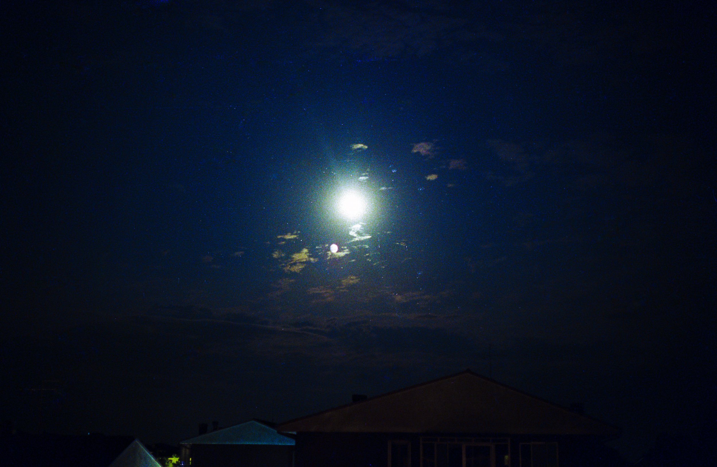 Night sky with bright moon, starry sky, and a few clouds, above the silhouette of houses.