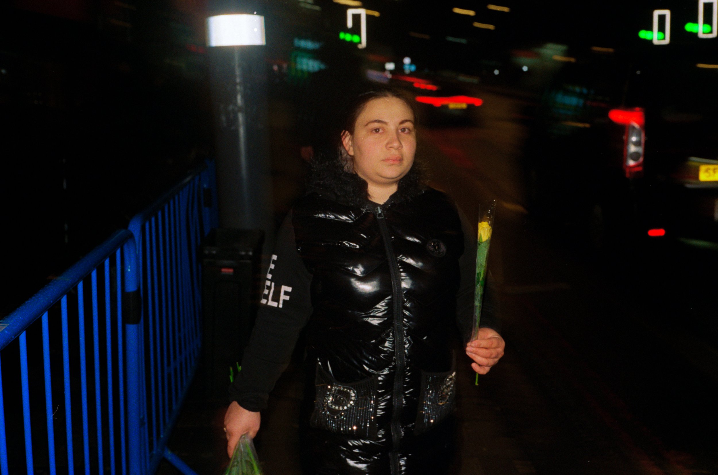A woman standing on a city sidewalk at night, holding flowers and a plastic bag, with cars and blurred city lights in the background.