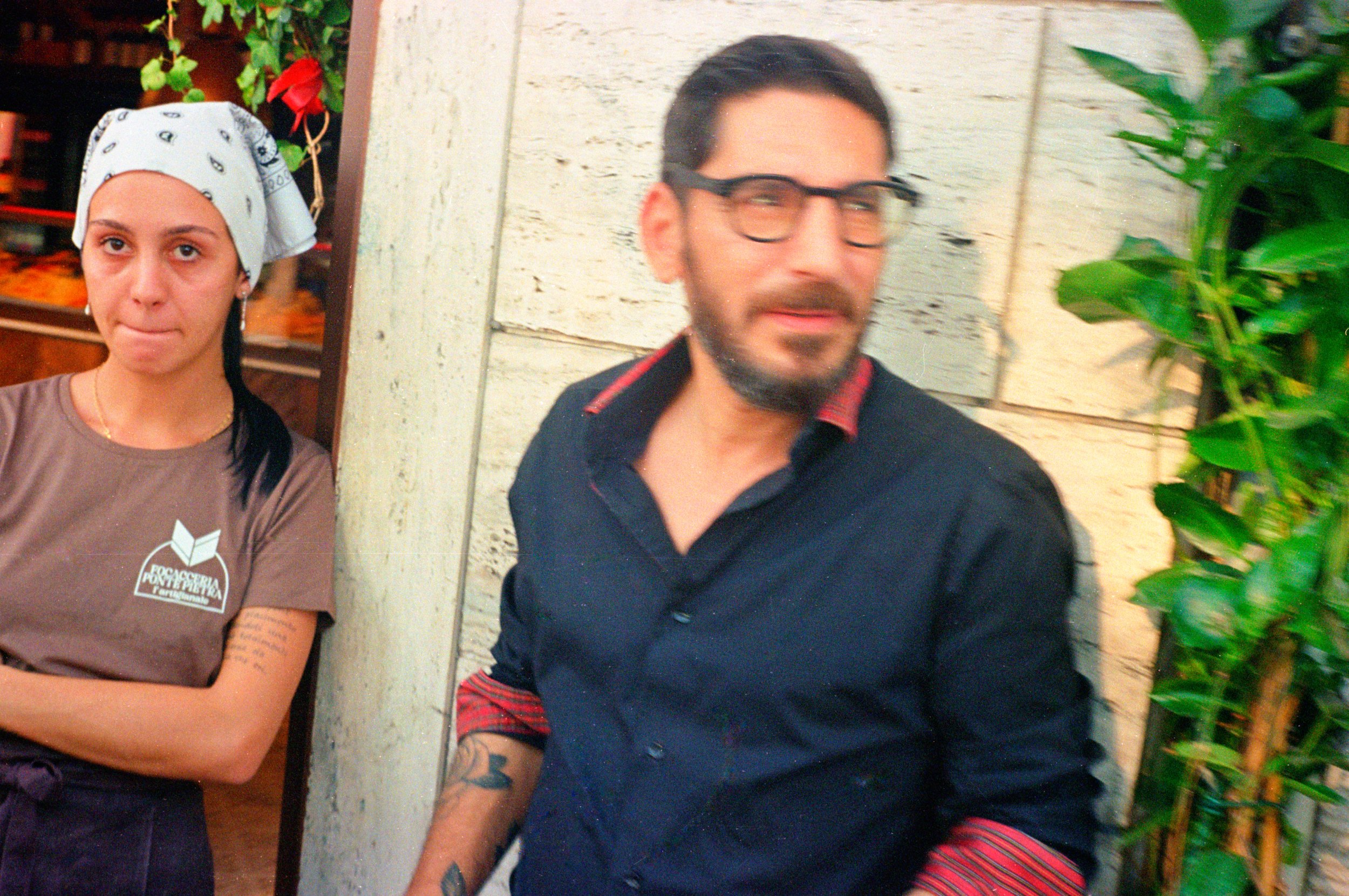 A woman wearing a white bandana and brown shirt standing next to a man with glasses and a dark shirt against a rustic background.