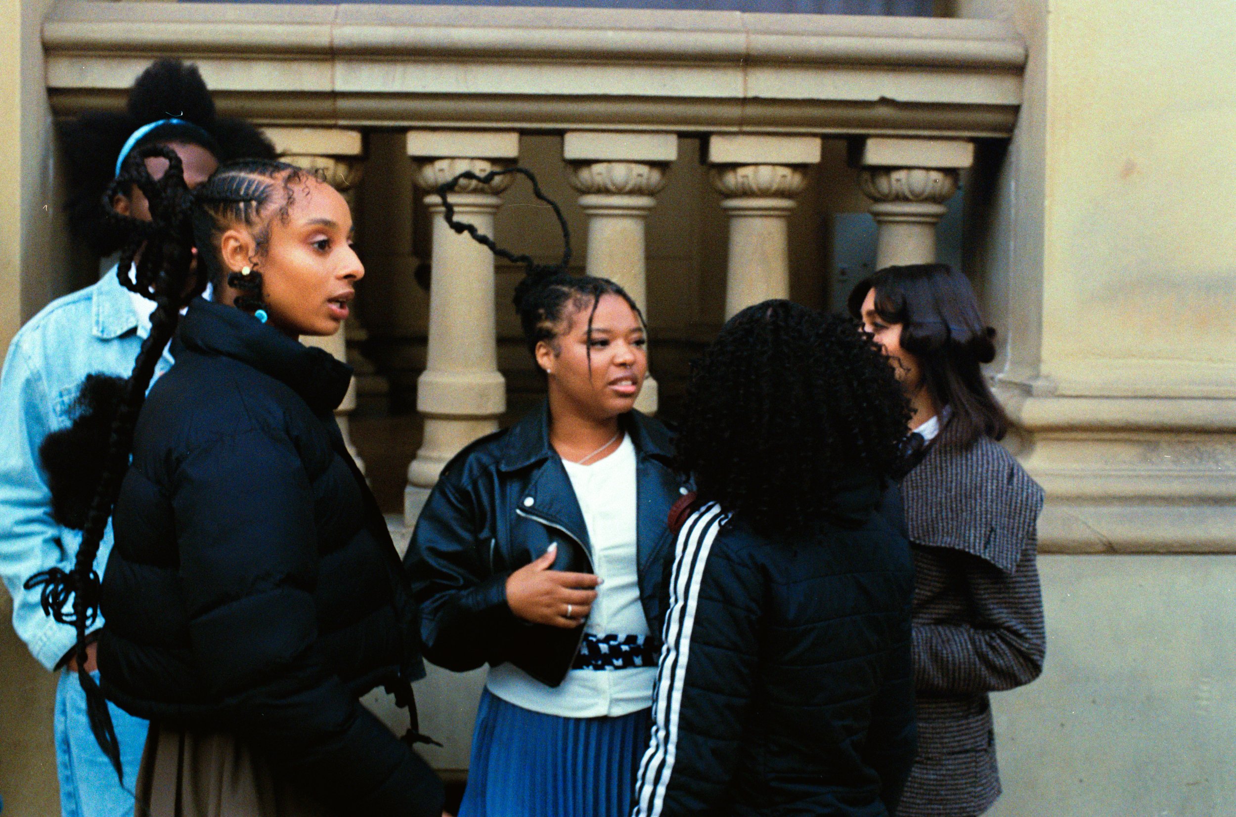 A group of five young women having a conversation indoors, standing near a staircase with decorative columns.