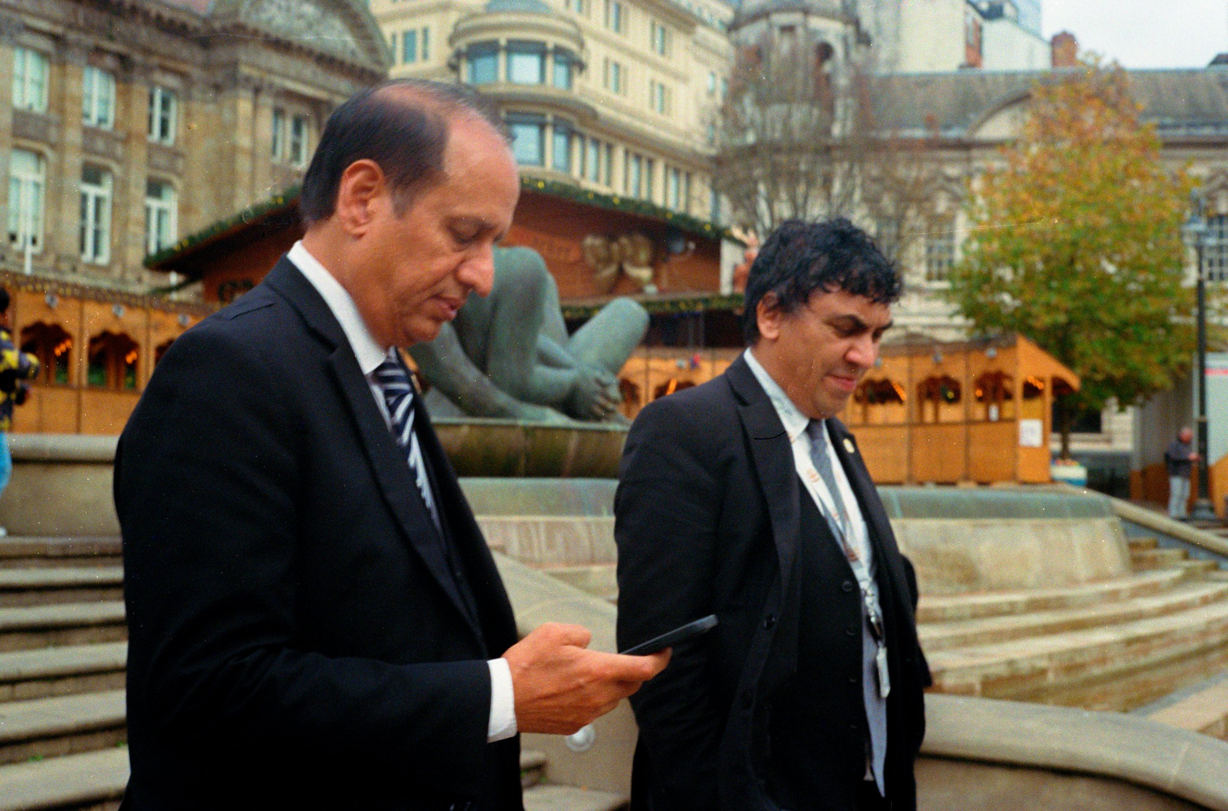 Two men in suits standing in front of a fountain with a sculpture, and small wooden market stalls in the background