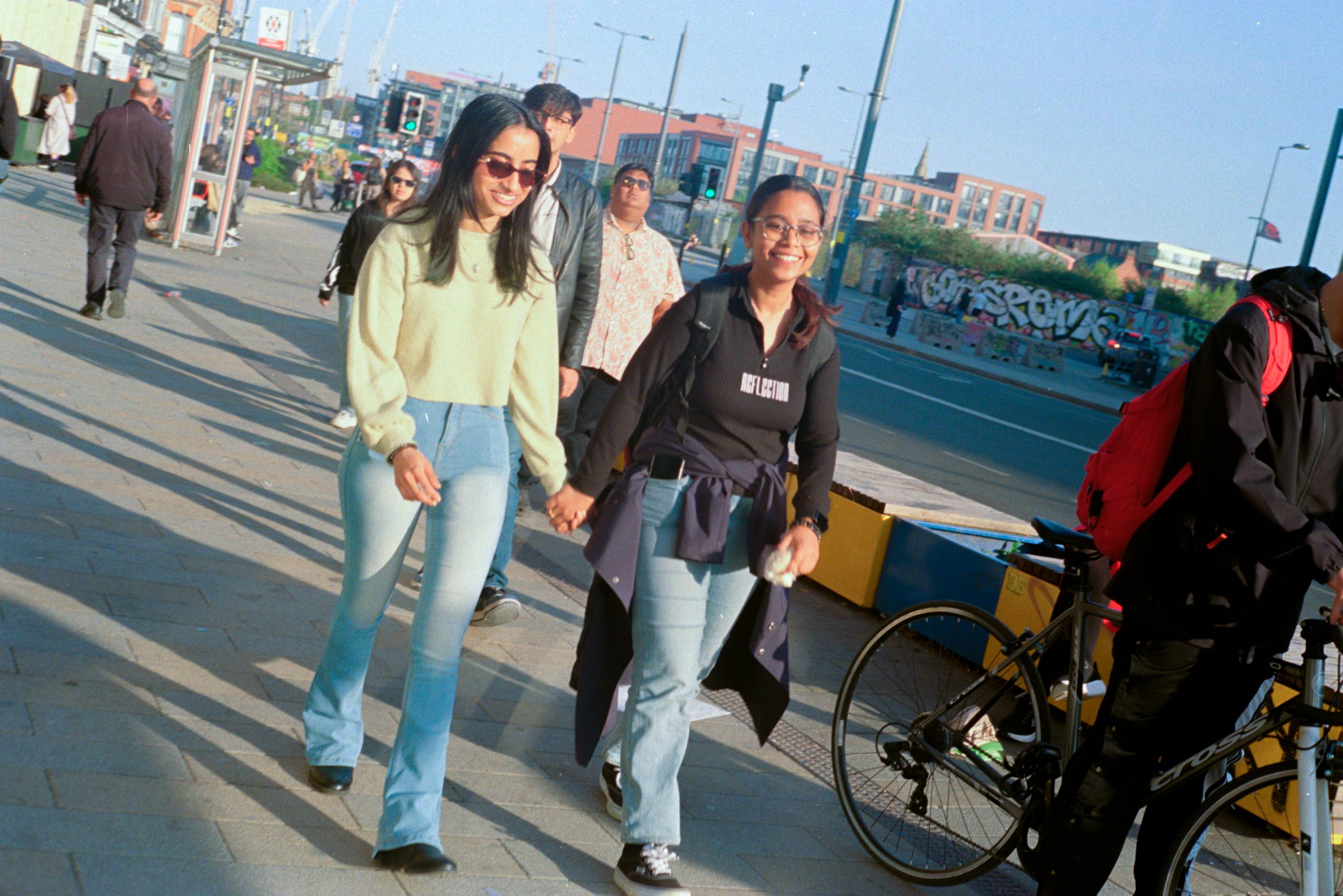 People walking on a city sidewalk during daytime, two women holding hands and smiling, with others in the background and colorful graffiti on a wall.