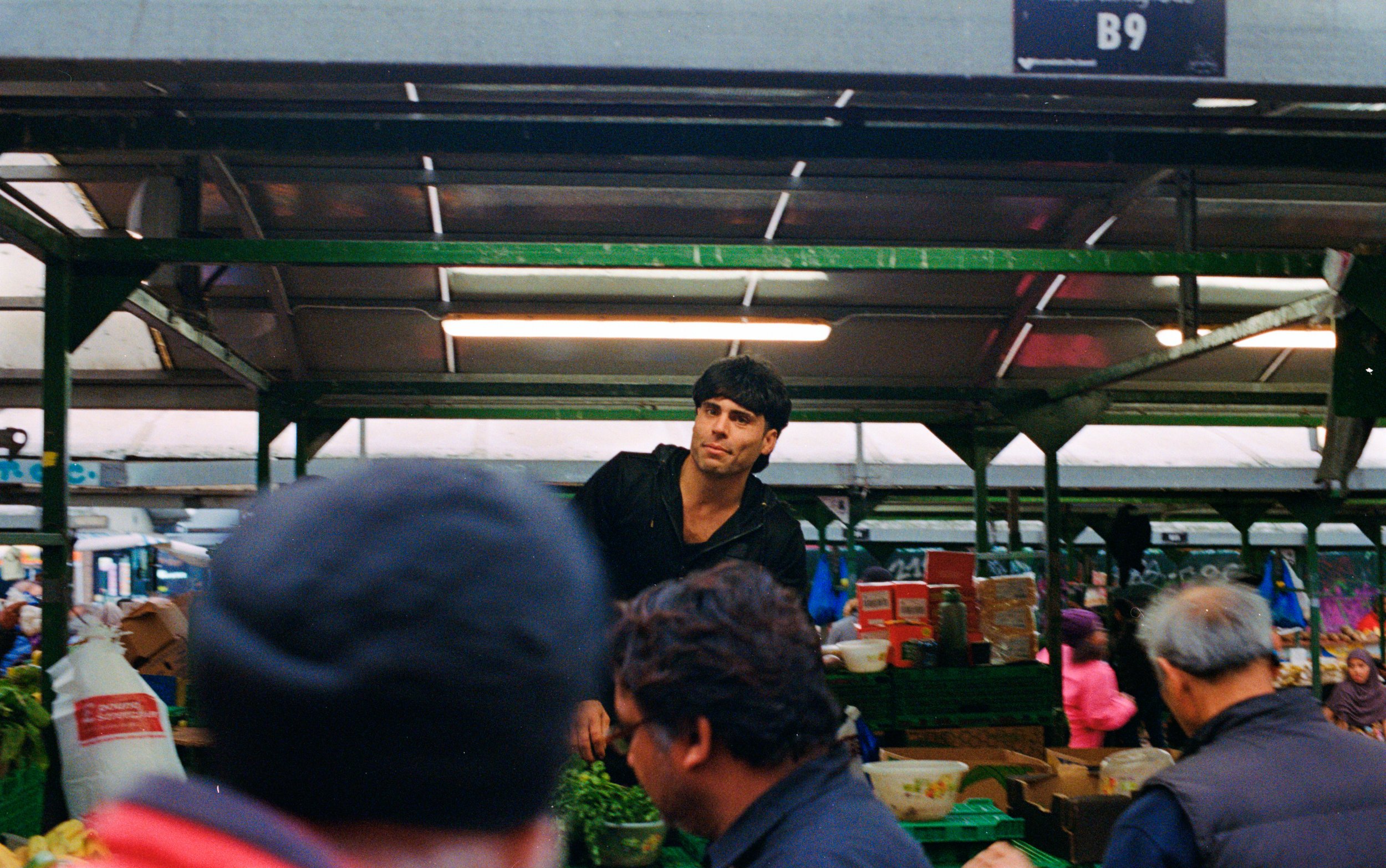 A man standing in a busy outdoor marketplace surrounded by people shopping and standing behind a produce stall with vegetables and packaged goods.
