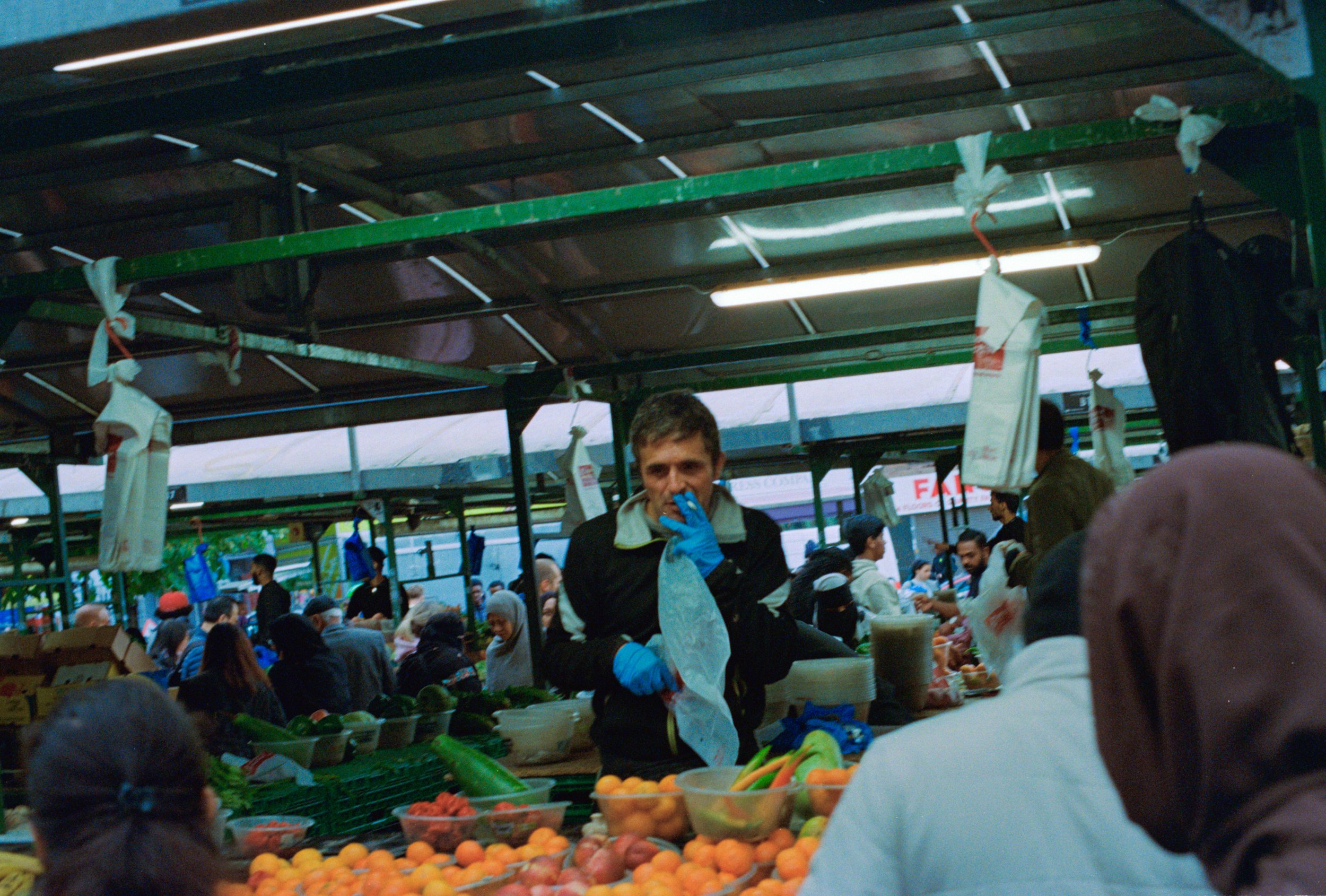A man wearing a black jacket and blue gloves is at a busy outdoor market stall, surrounded by fresh fruits and vegetables, with many people shopping in the background under a green canopy.
