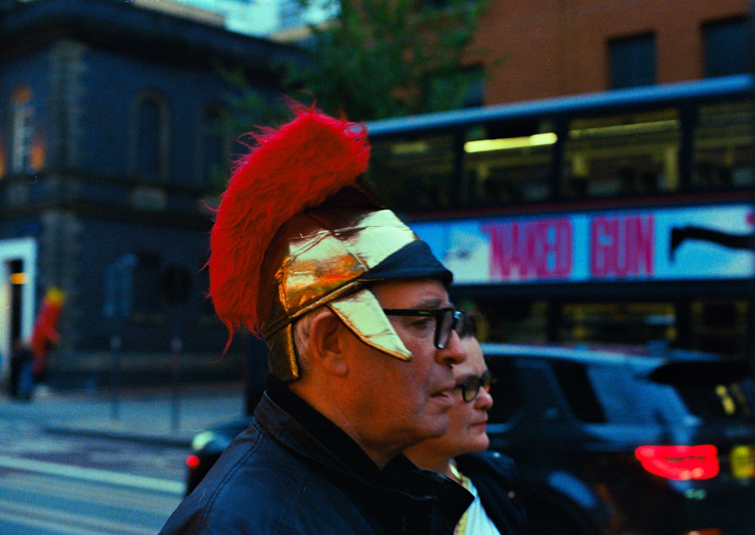 A man wearing a shiny gold and black helmet with a red feather crest and black glasses, standing on a city street during twilight. There is a large double-decker bus and other cars in the background.