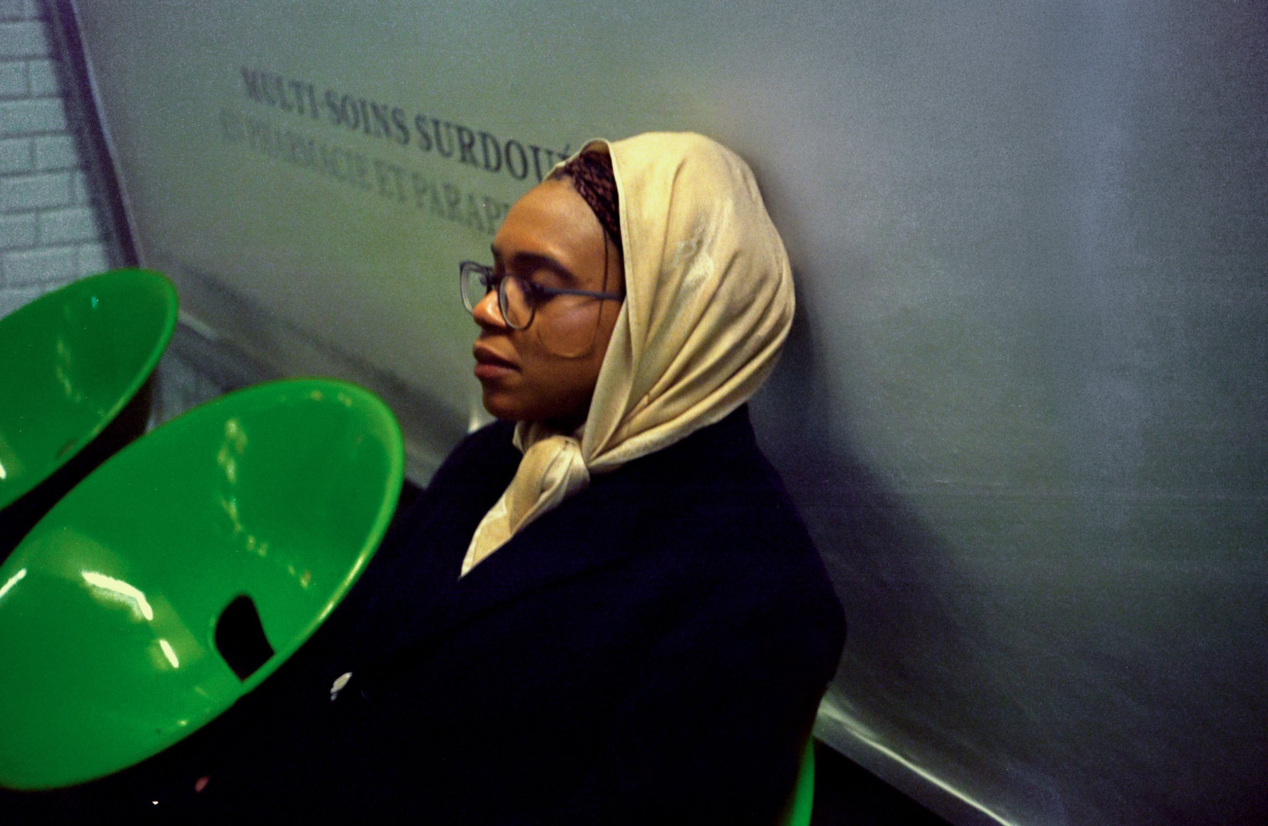A woman with glasses and a beige headscarf sitting against a wall with green chairs around her.