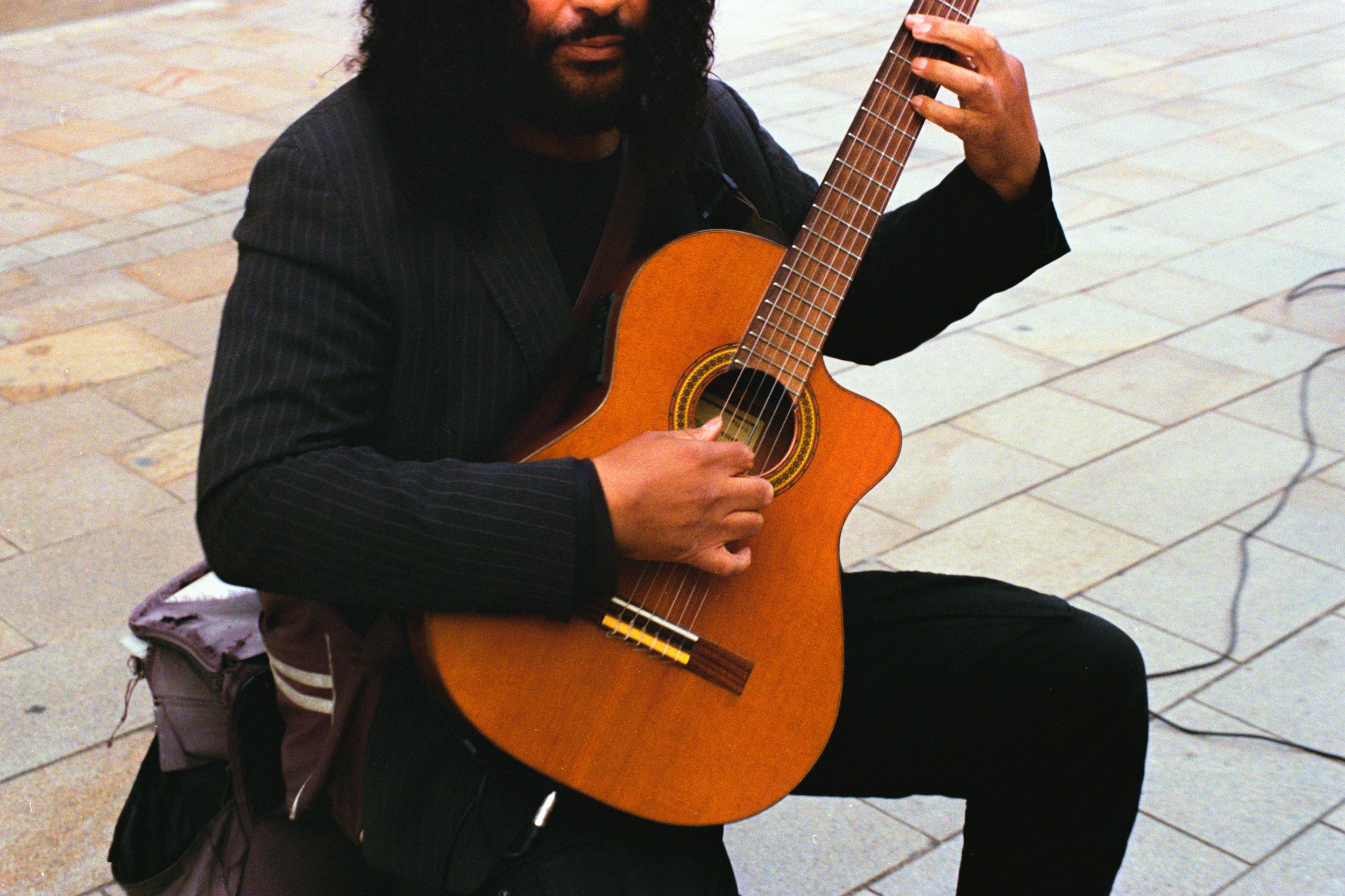 A man with long black curly hair and beard is playing an acoustic guitar while sitting on a sidewalk.