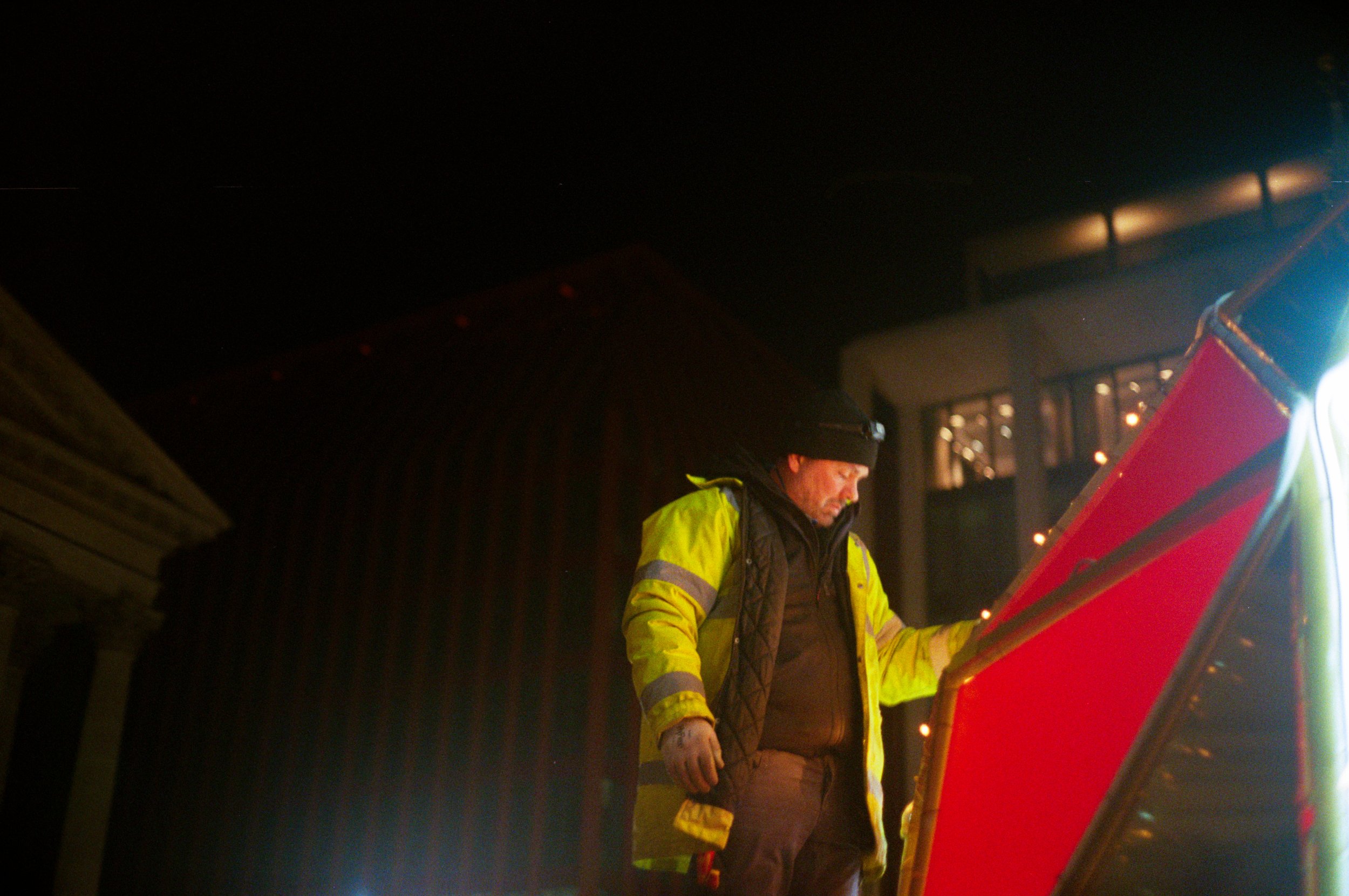 A man in a yellow high-visibility jacket and black beanie hat standing outside at night, looking at a red object.