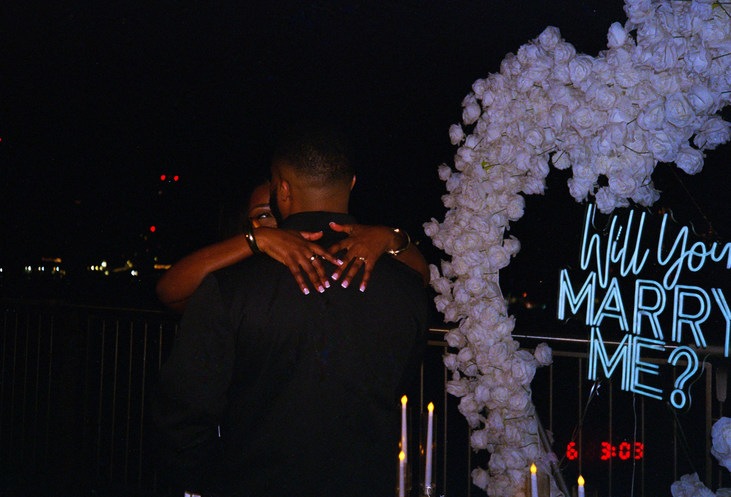 A couple embracing at night on a rooftop, with a decorative floral arch and a neon sign that reads 'Will you marry me?' in the background.