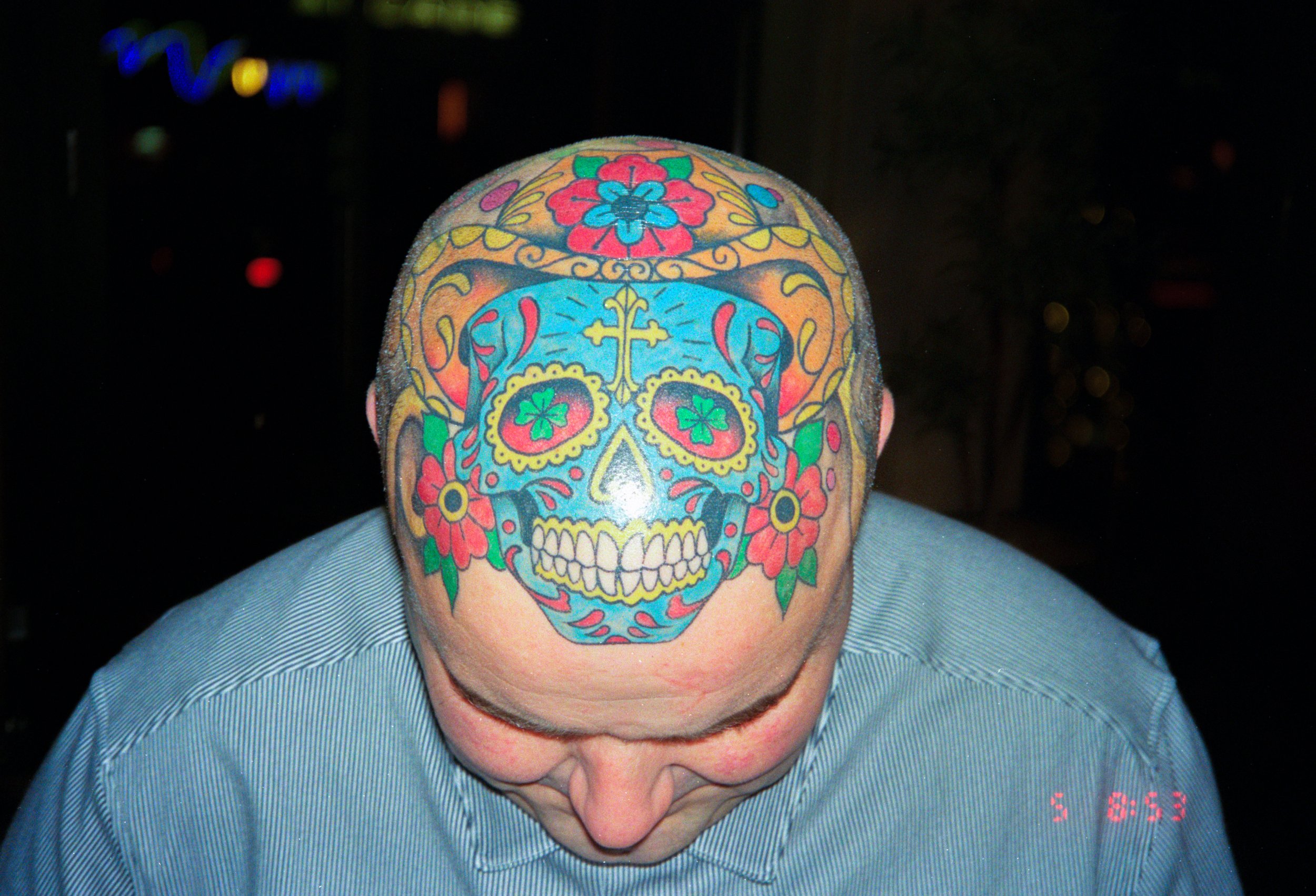 A person with a colorful sugar skull tattoo on their bald head, wearing a light blue shirt, looking downward in a dark background.