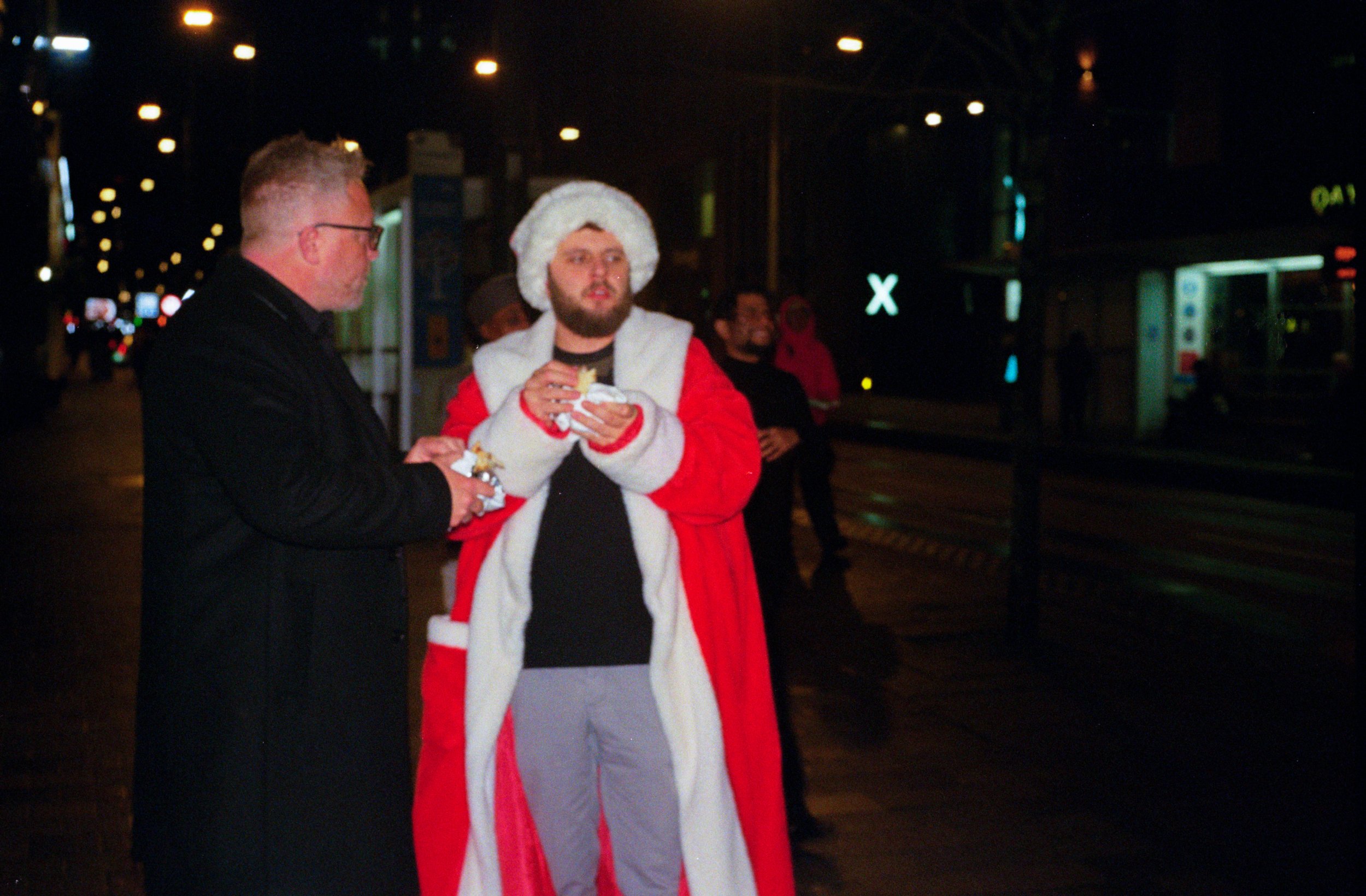 People on a city street at night, one dressed as Santa Claus with a red coat and hat, holding food, others dressed in dark clothing, some in the background.