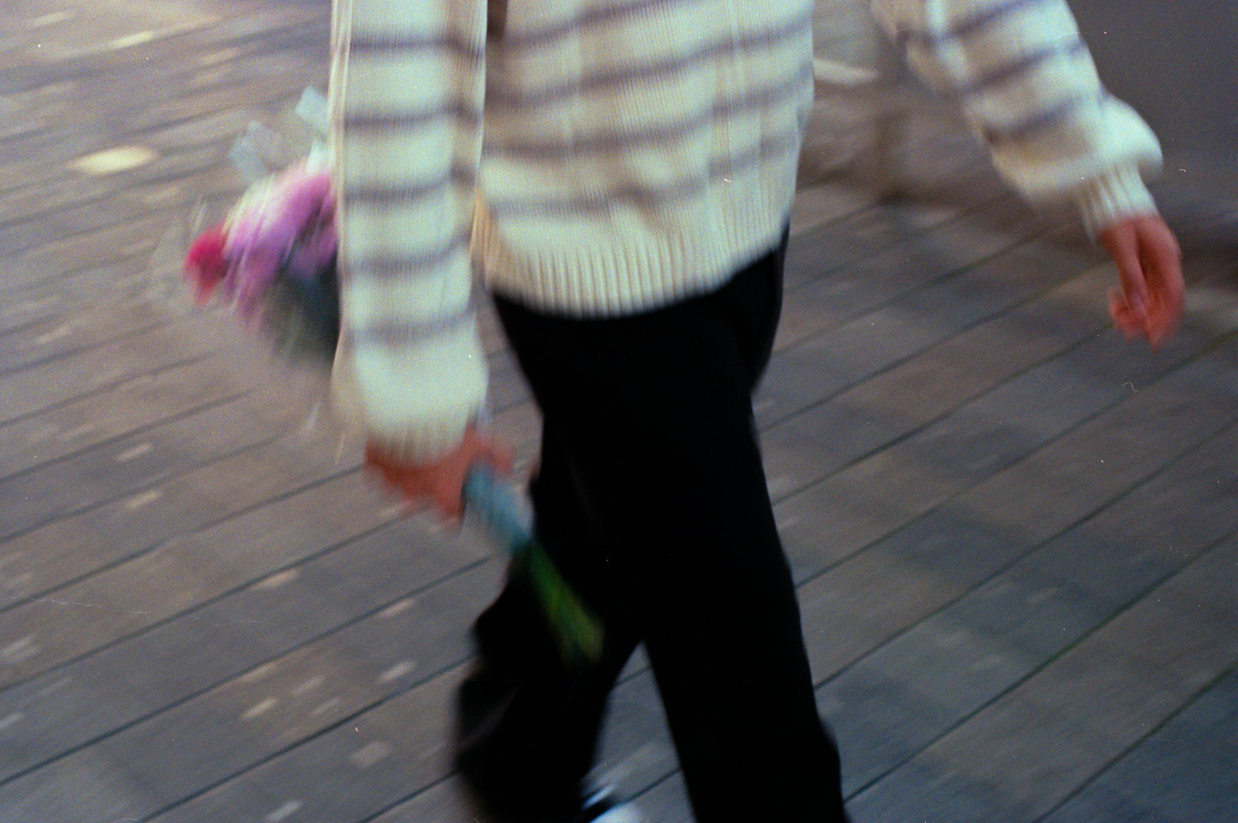 A person wearing a light-colored striped sweater and black pants walking on a wooden boardwalk, holding a bouquet of pink and purple flowers in their left hand.
