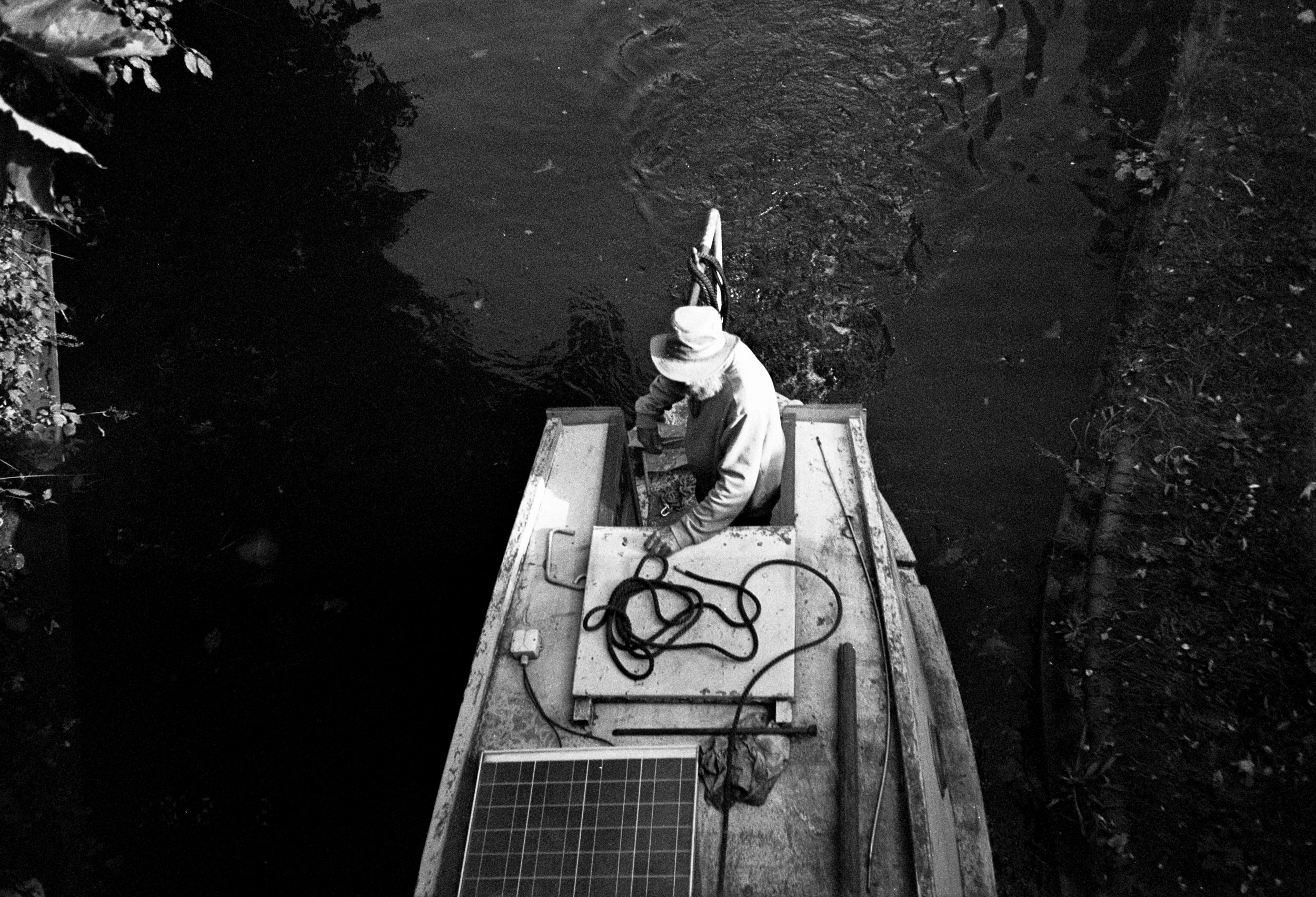 A person is standing on the deck of a boat, tying a rope to the railing, with water below and surrounding vegetation visible on the riverbank.